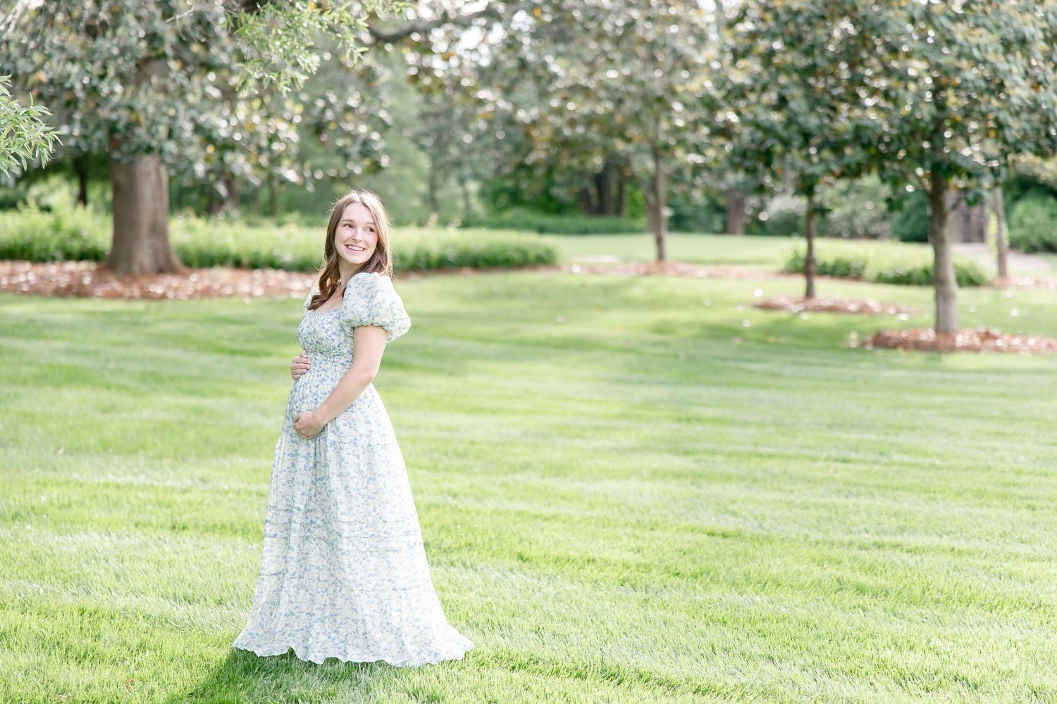 A figure in a flowing white lace maternity dress poses gracefully in a sunny park setting among trees and green grass.