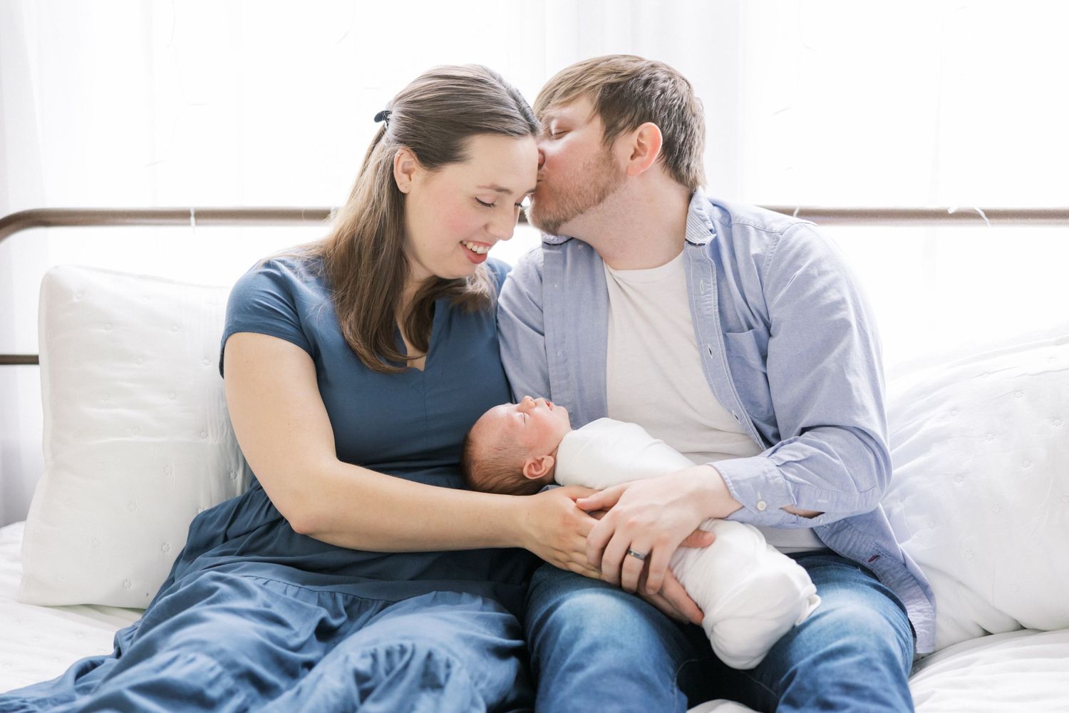 Parents cuddling with newborn on white bedding in intimate setting.