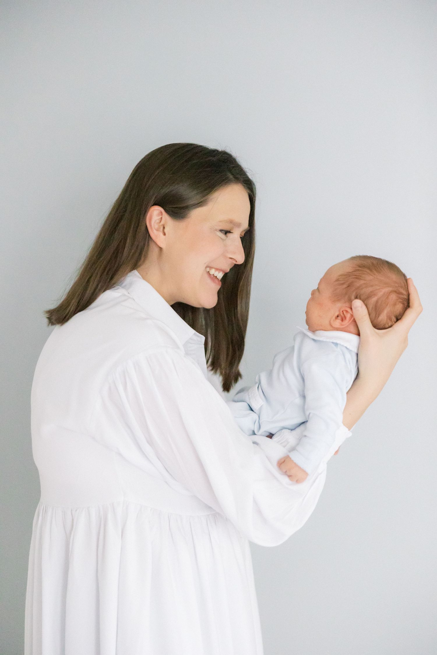 A woman in white dress gazes lovingly at her newborn baby against a light gray background.