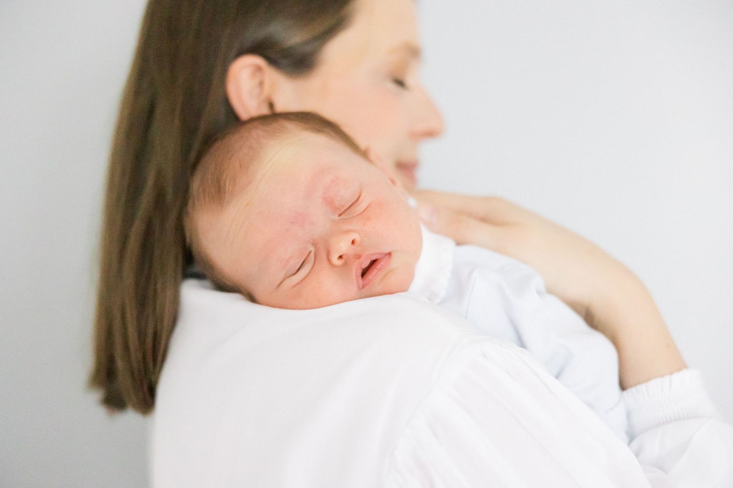 Sleeping newborn baby wearing white rests peacefully against shoulder in soft natural light.