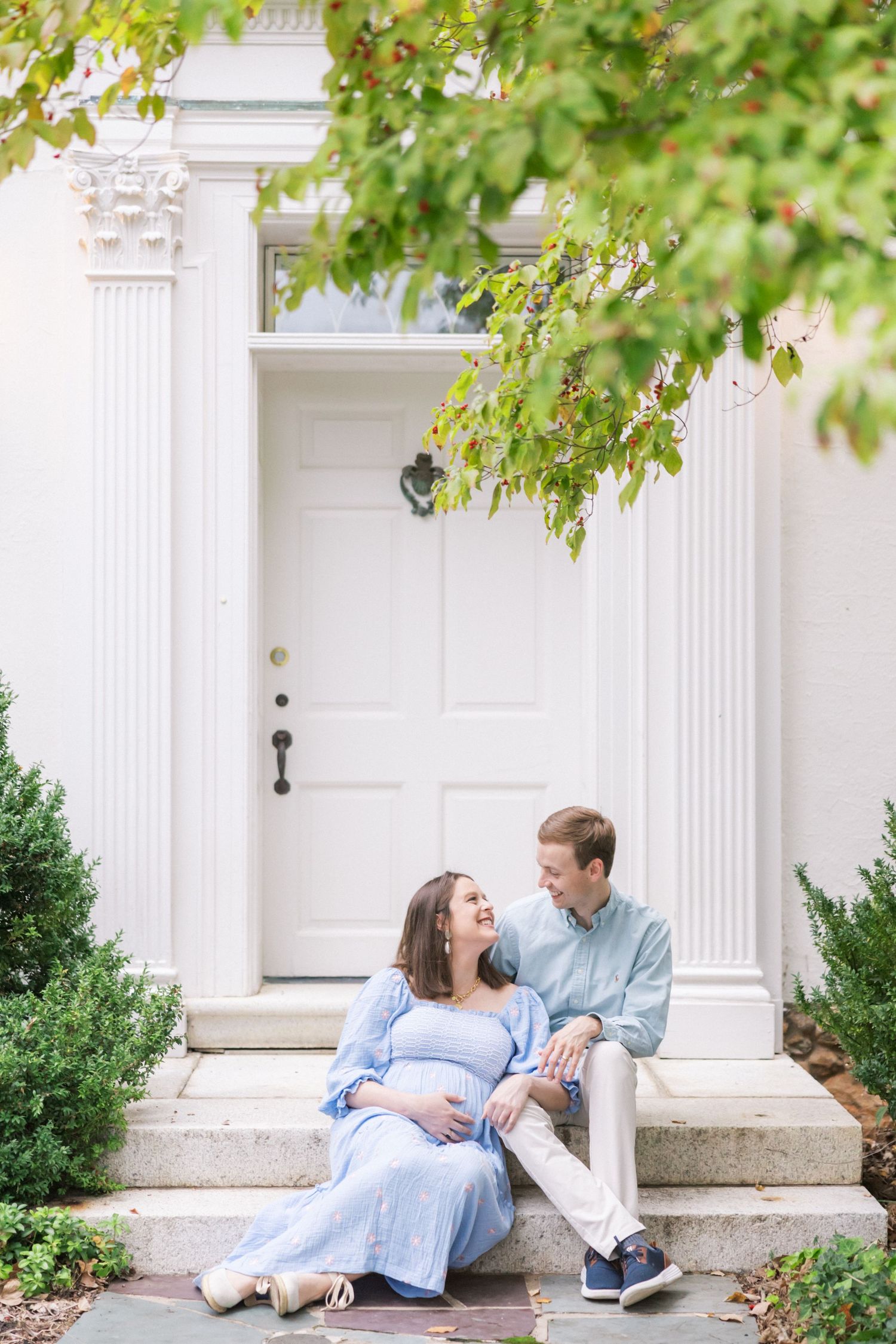 Expecting couple sits on white columned doorstep surrounded by greenery in a romantic maternity portrait.