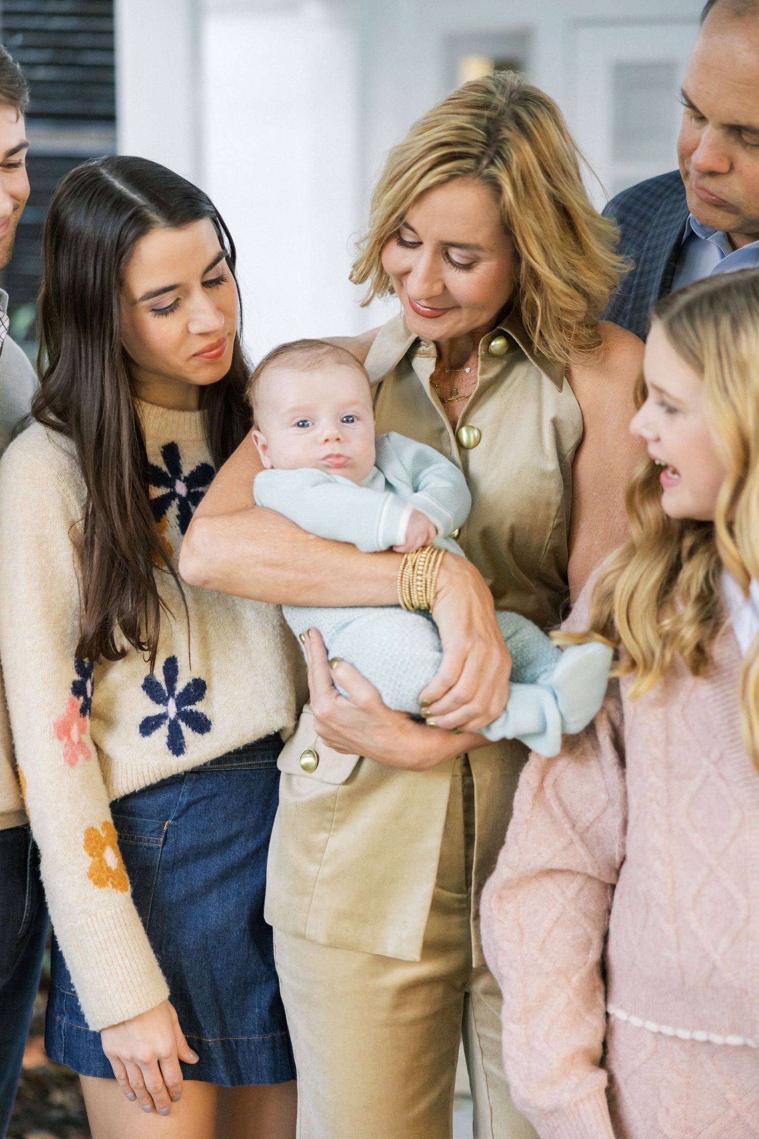 Family members gathered indoors admiring a baby in casual neutral-toned clothing.