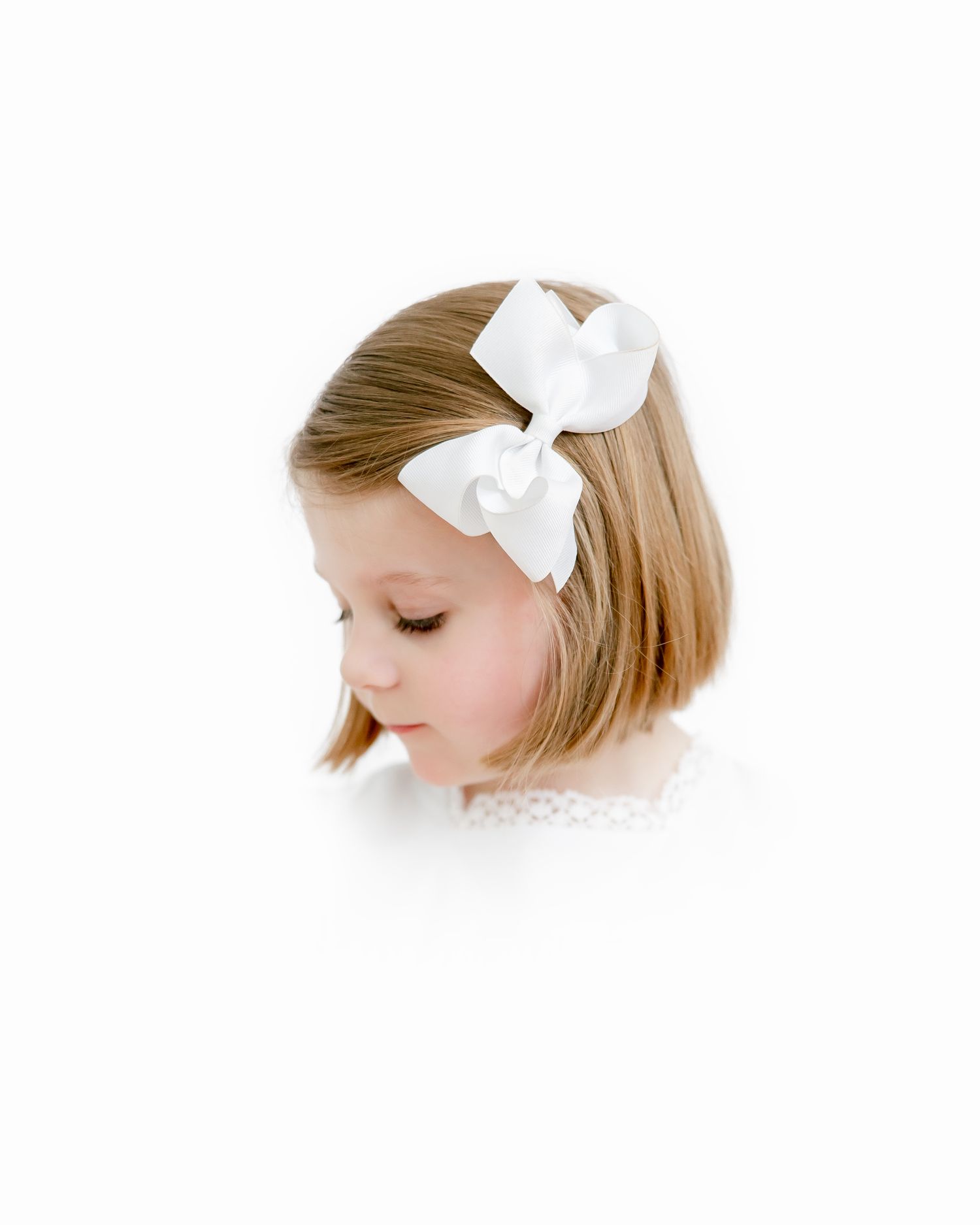 A young child with a short bob haircut wearing a white bow accessory looks down pensively against a white background.