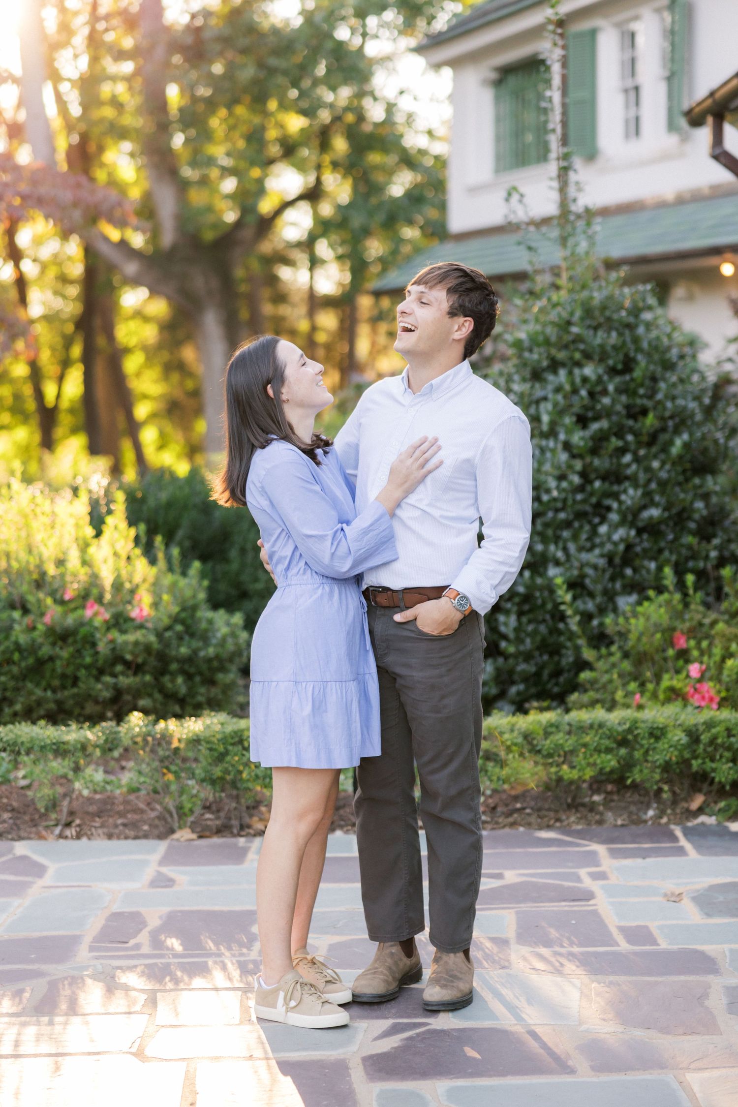 A romantic couple embraces on a stone patio during golden hour with a house and garden in the background.
