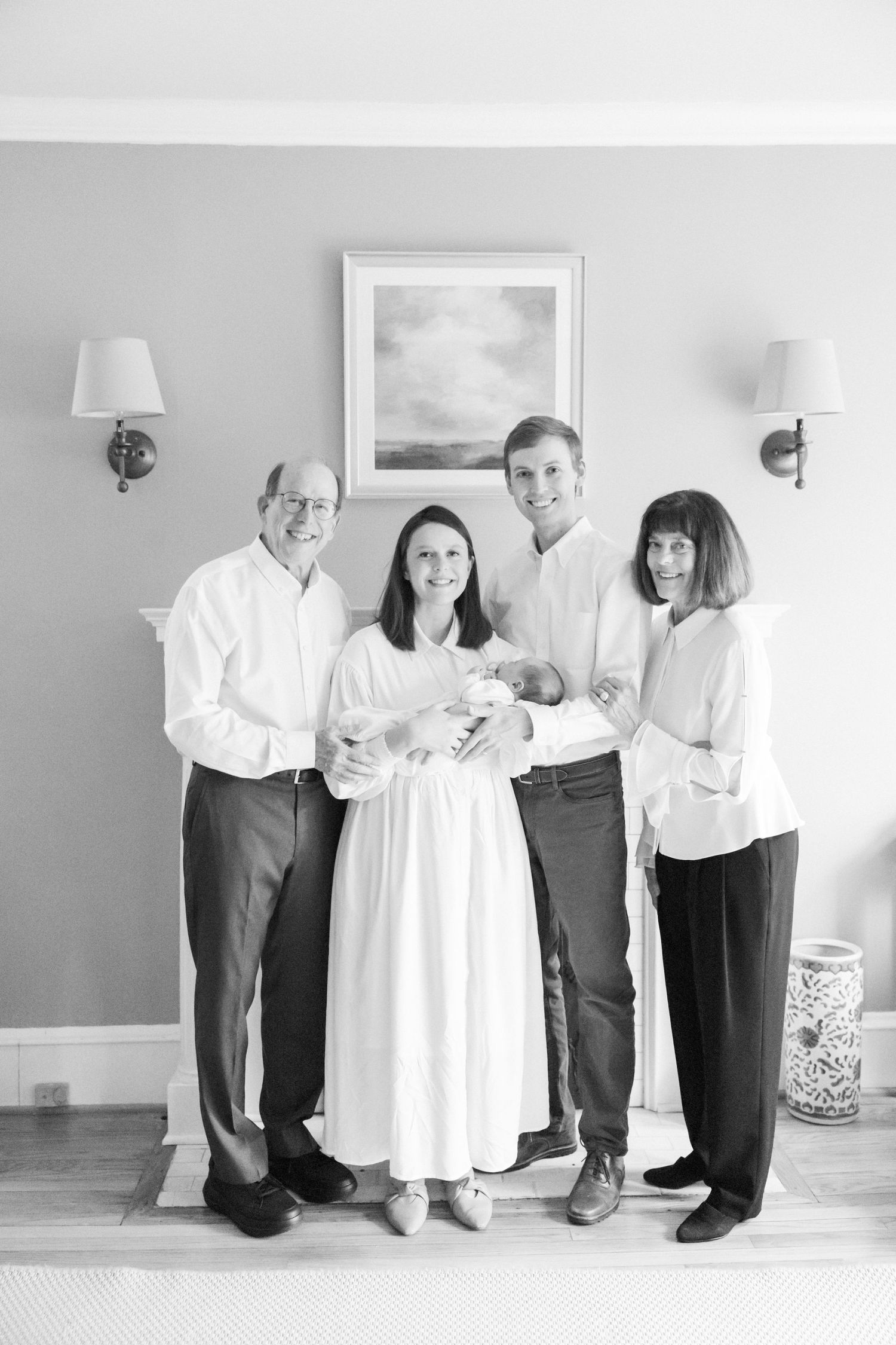 A family portrait with a newborn baby taken in front of a gray wall with artwork and sconces.