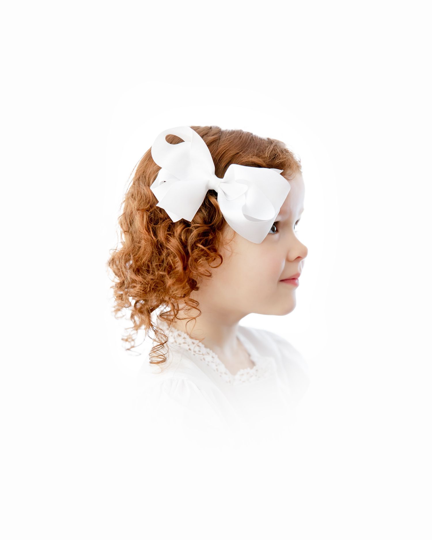 Side profile portrait of a child with curly red hair wearing a white bow and white top.