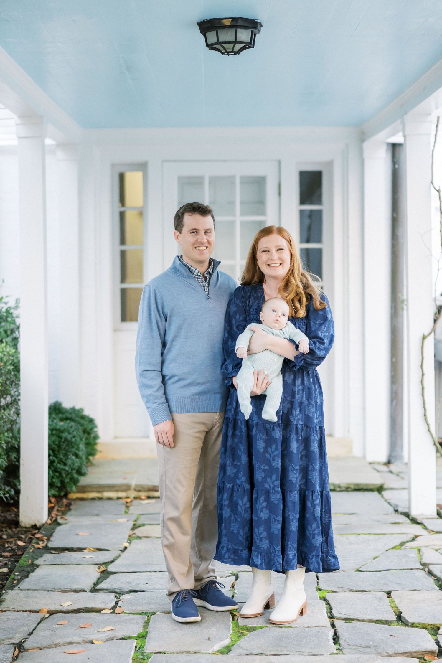 A couple stands on a white porch with their newborn baby on a bright sunny day.