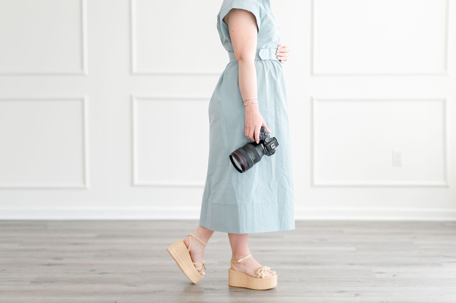 Person in a light blue dress holding a camera while wearing beige platform sandals stands against a white wall.