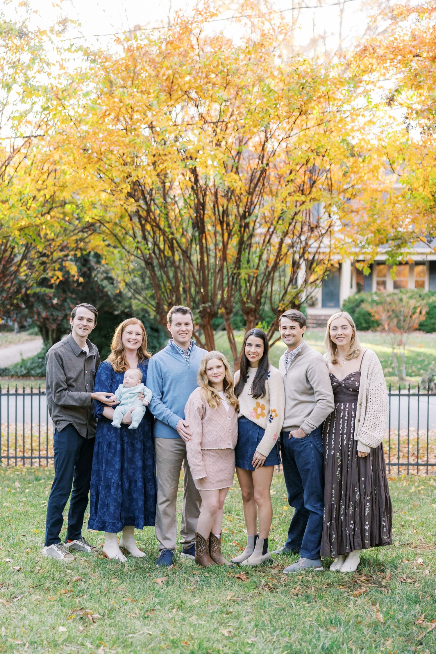 Family group portrait outdoors in fall with golden trees and iron fence in background.
