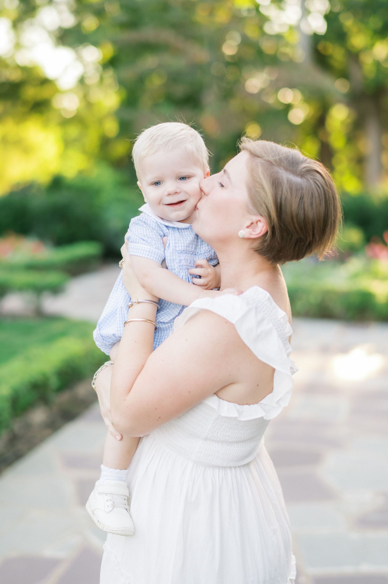 Mother in white dress kisses baby in outdoor garden setting with natural bokeh background.