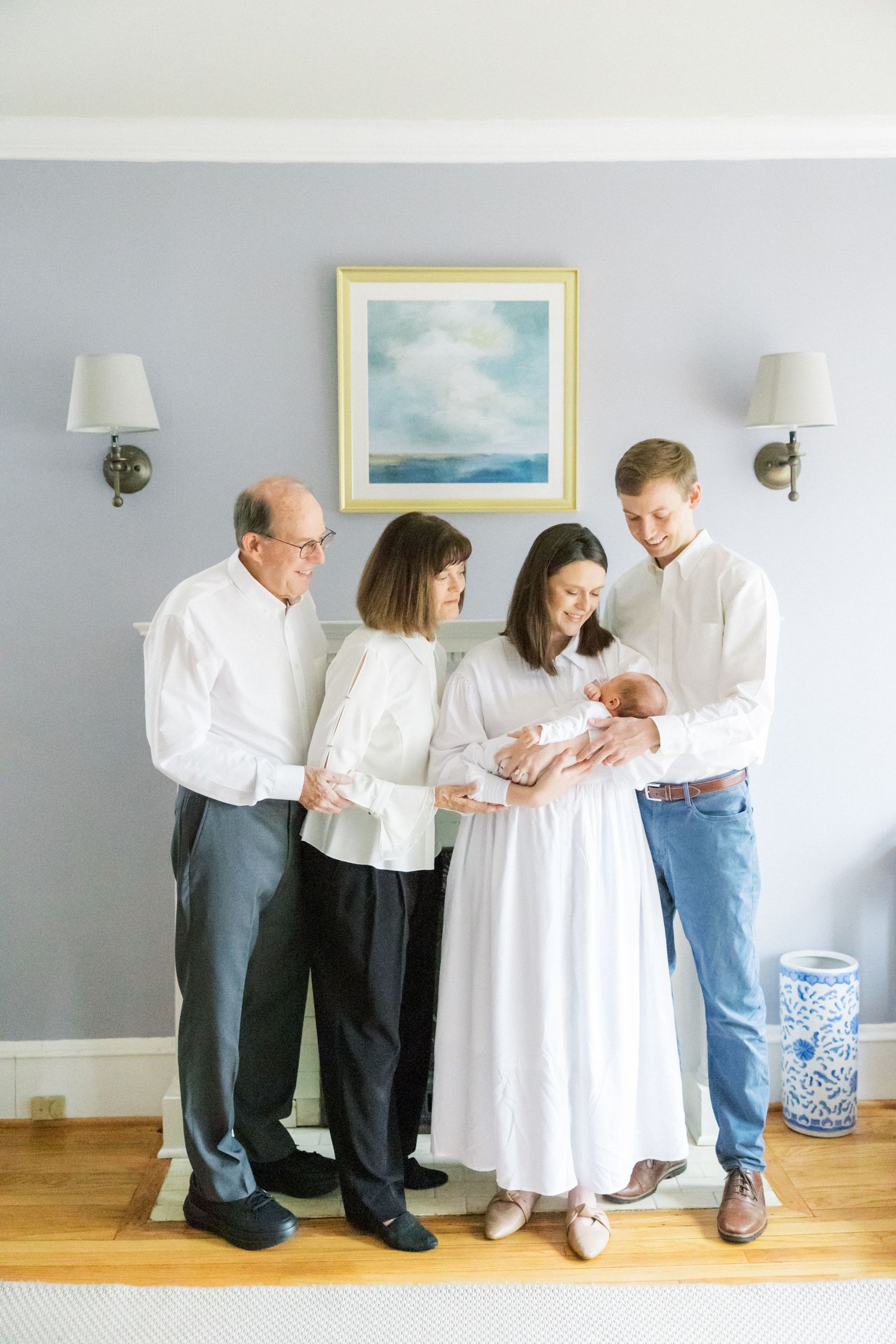 A family gathering indoors admiring a newborn baby against a grey wall with a seascape painting and wall sconces.