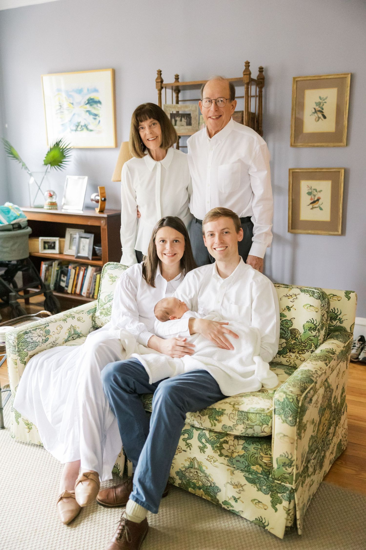 Multi-generational family wearing white sits together on floral couch with newborn baby.