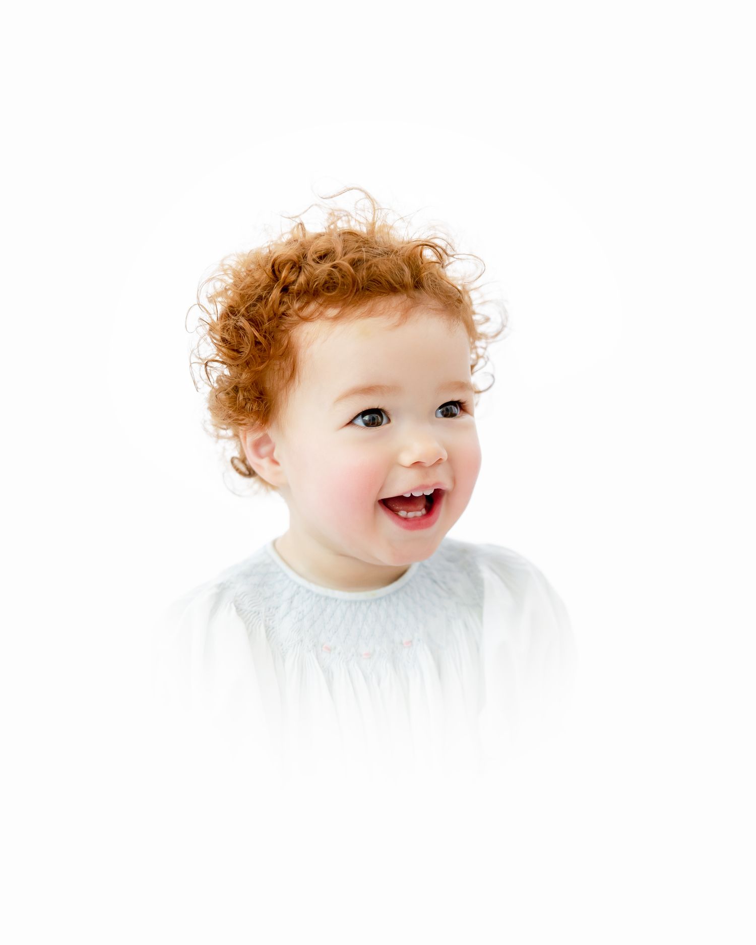 Joyful toddler with curly ginger hair wearing a white shirt against a white background.