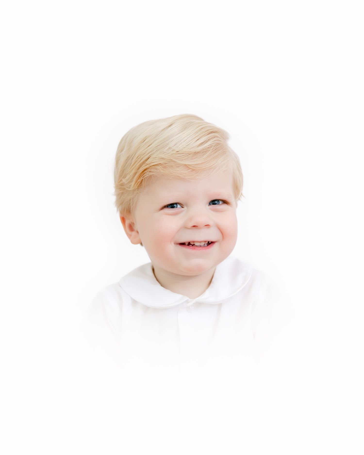 A smiling toddler wearing a white shirt poses for a studio portrait against a white background.