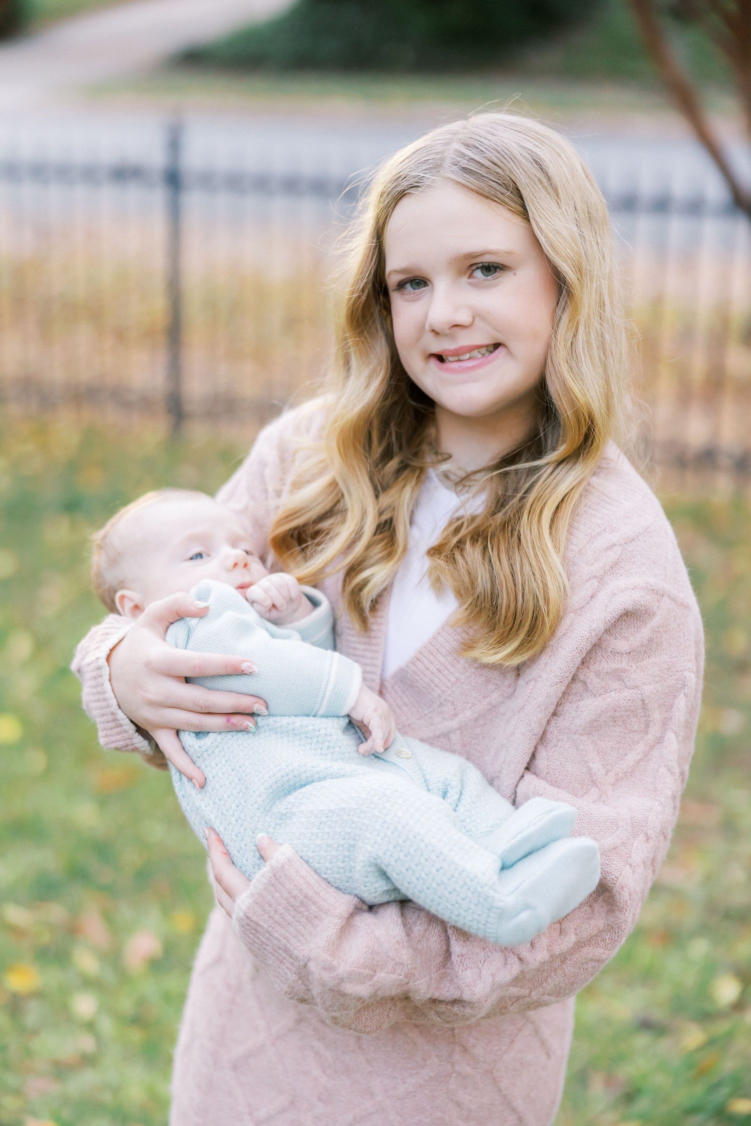 Young girl in pink sweater holding a baby in light blue outfit against fall foliage background.