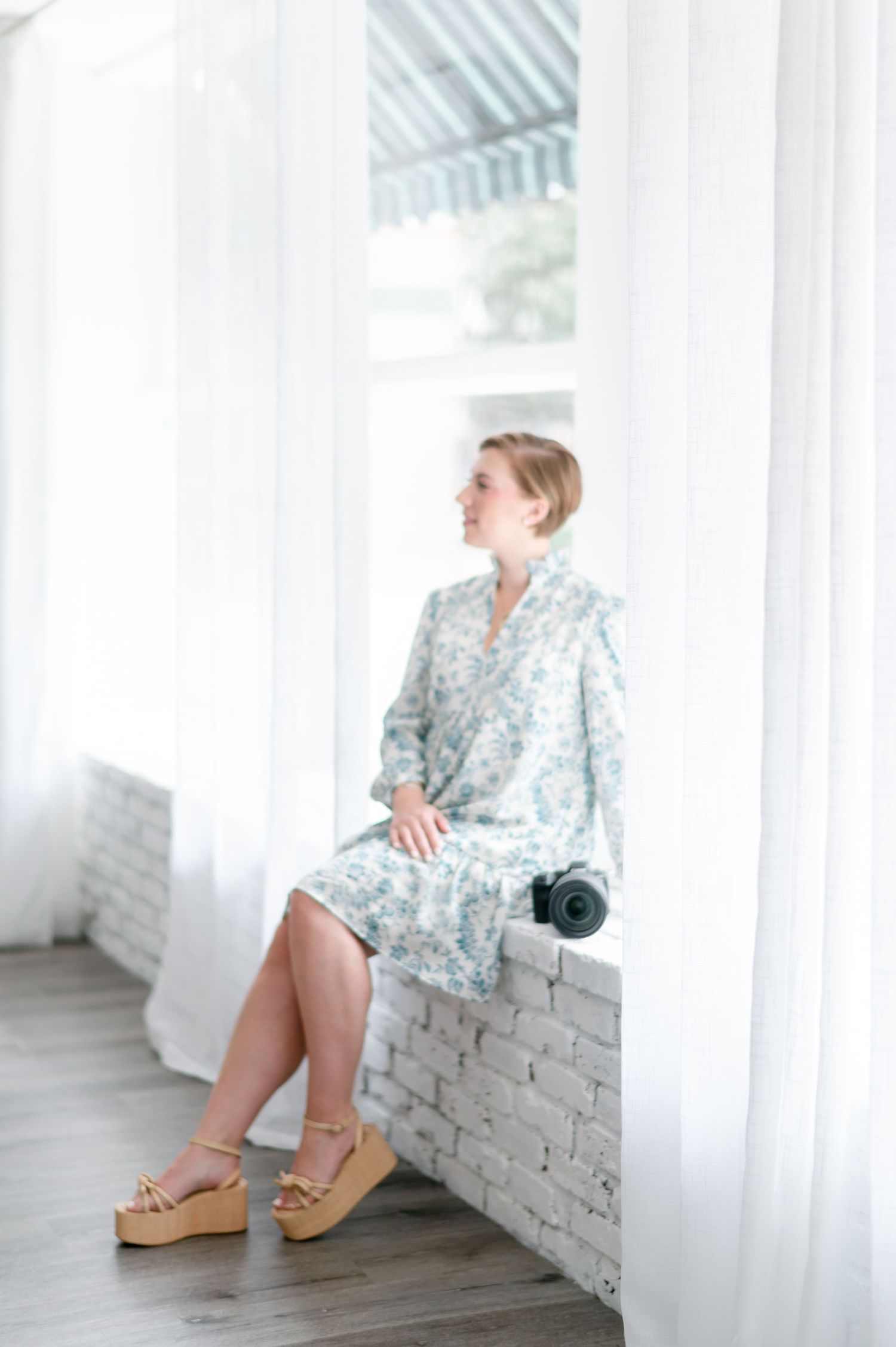 Professional photographer in a floral dress sits on white brick ledge with camera.