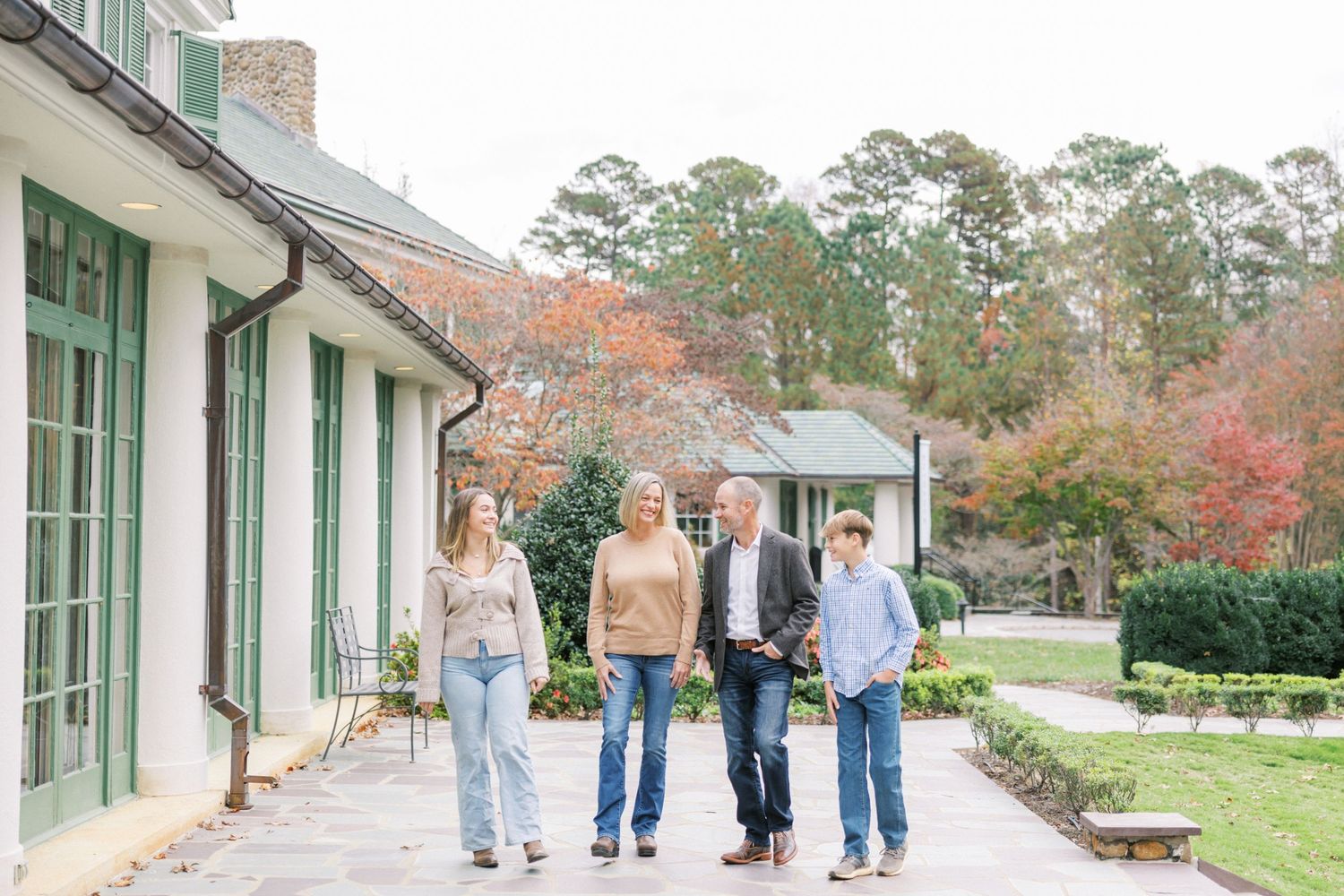 Four people walking together on a paved path near a white building with autumn trees in the background.