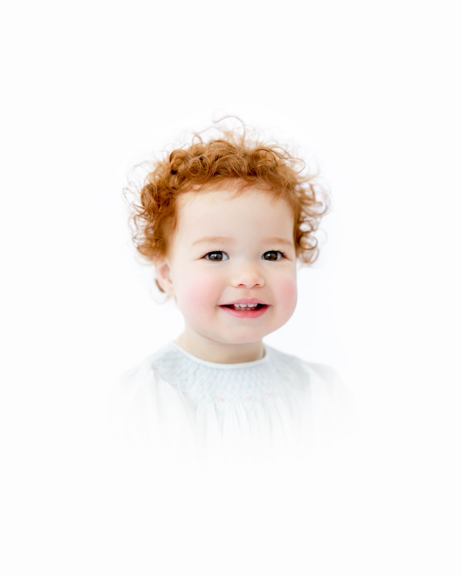 A sweet toddler with curly ginger hair and wearing white smiles brightly against a white background.