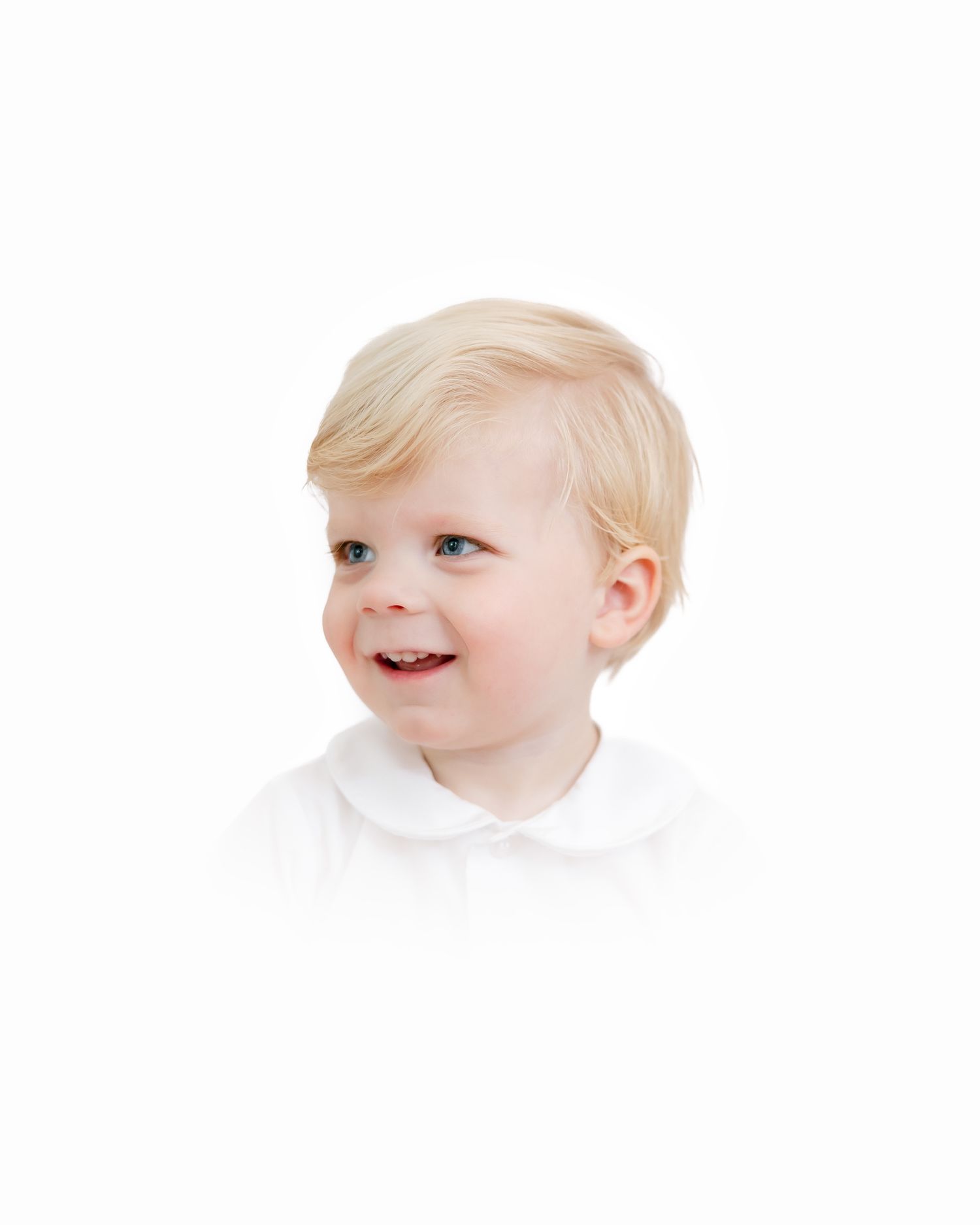 A small child with light blonde hair wearing a white collared shirt smiles while looking to the side against a white background.