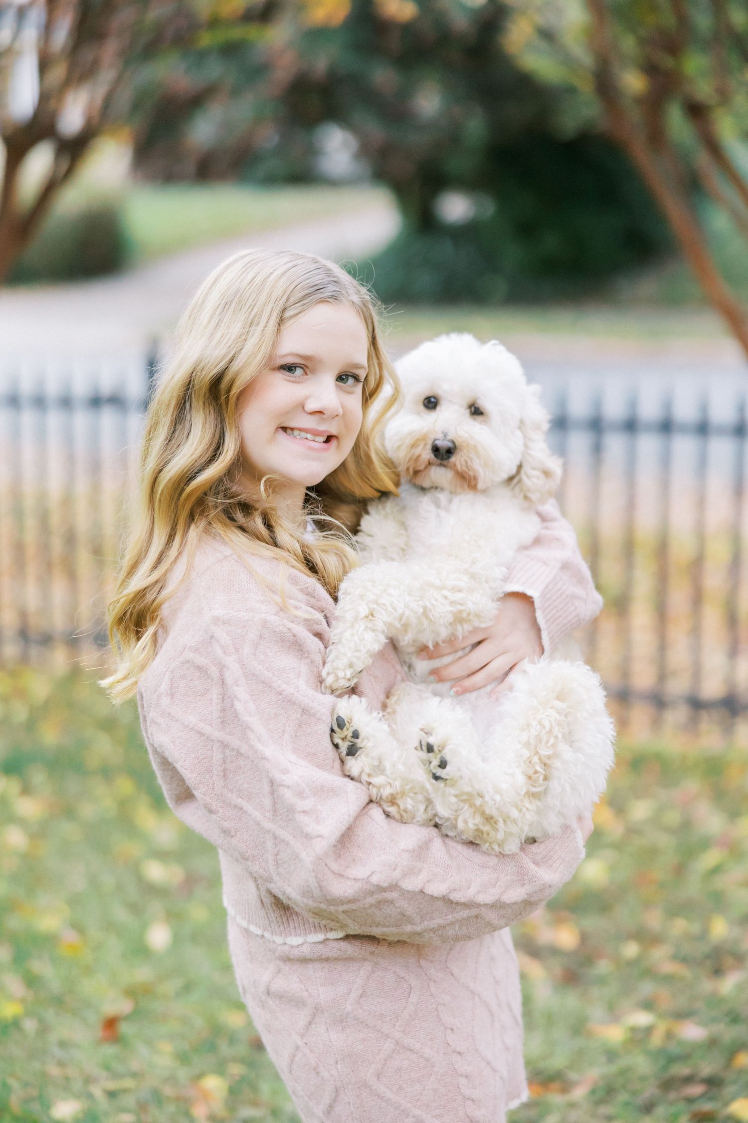Young person in a pink sweater holding a small white fluffy dog while standing in a fall outdoor setting.