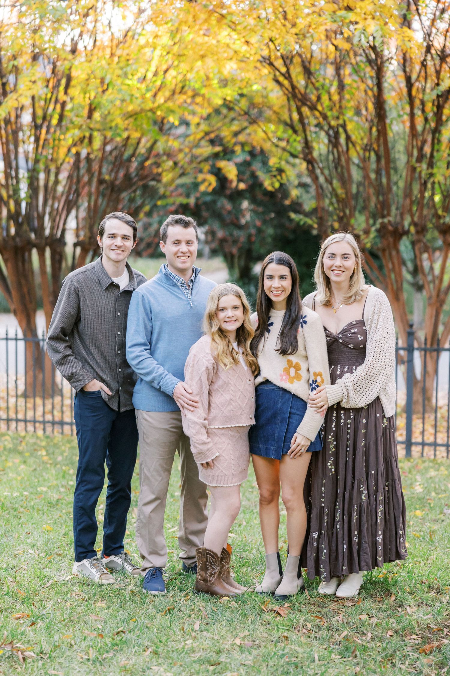 Five people pose together for a family portrait in front of autumn trees with golden leaves.