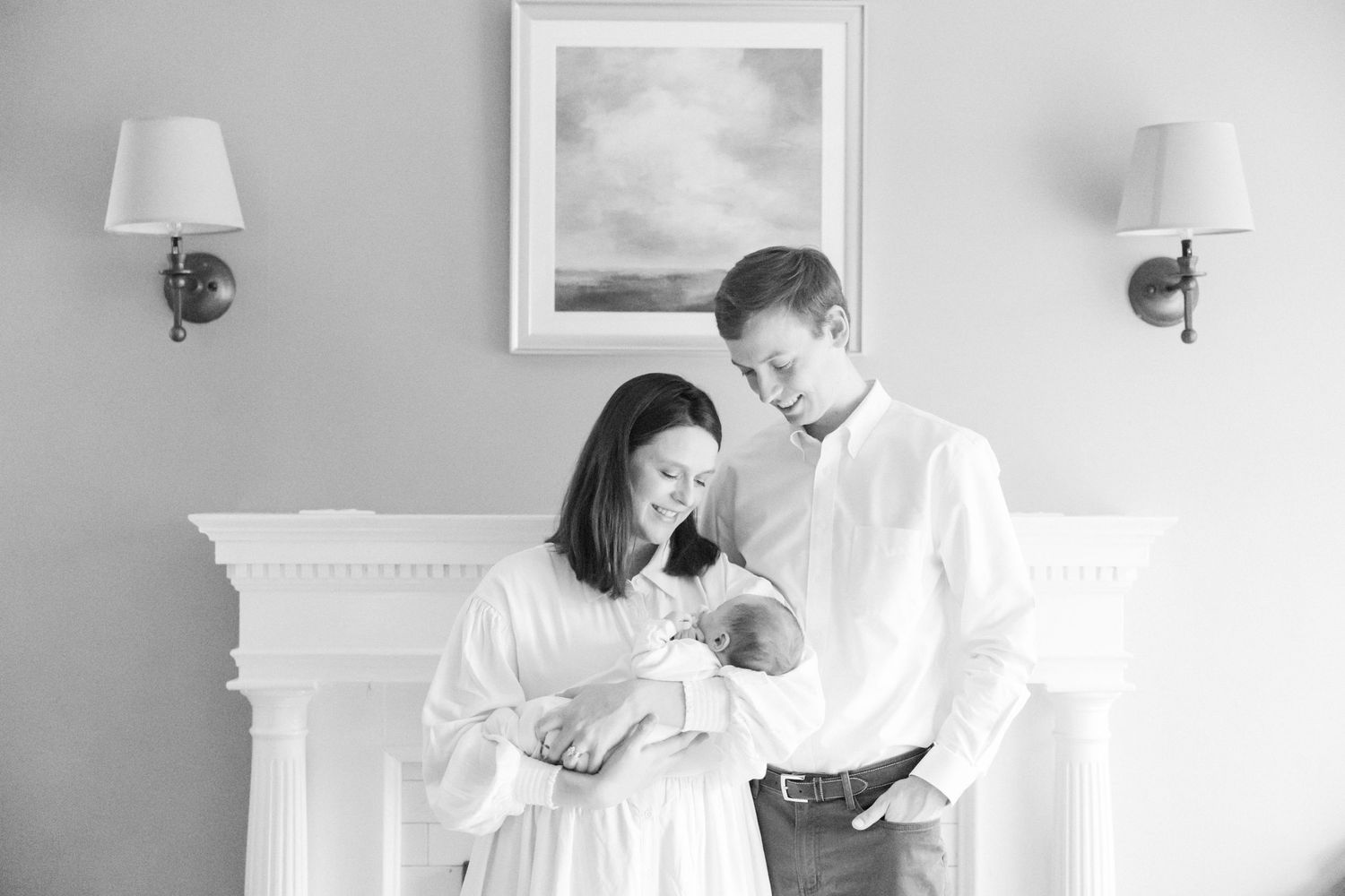 A tender moment in black and white with a couple and newborn baby standing near a mantle with wall sconces.