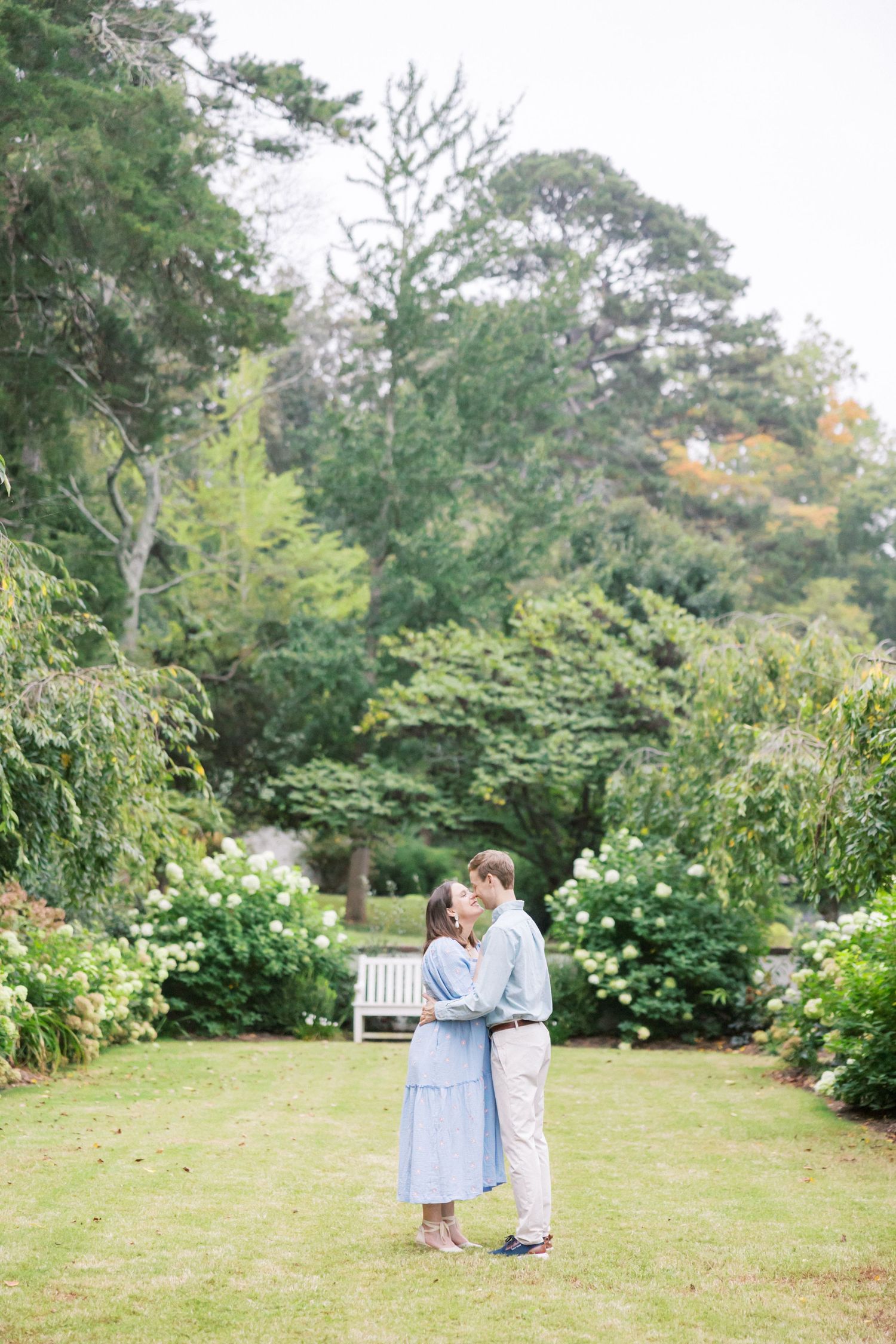 Couple embraces on a lush garden lawn surrounded by blooming bushes and tall trees during maternity photoshoot.