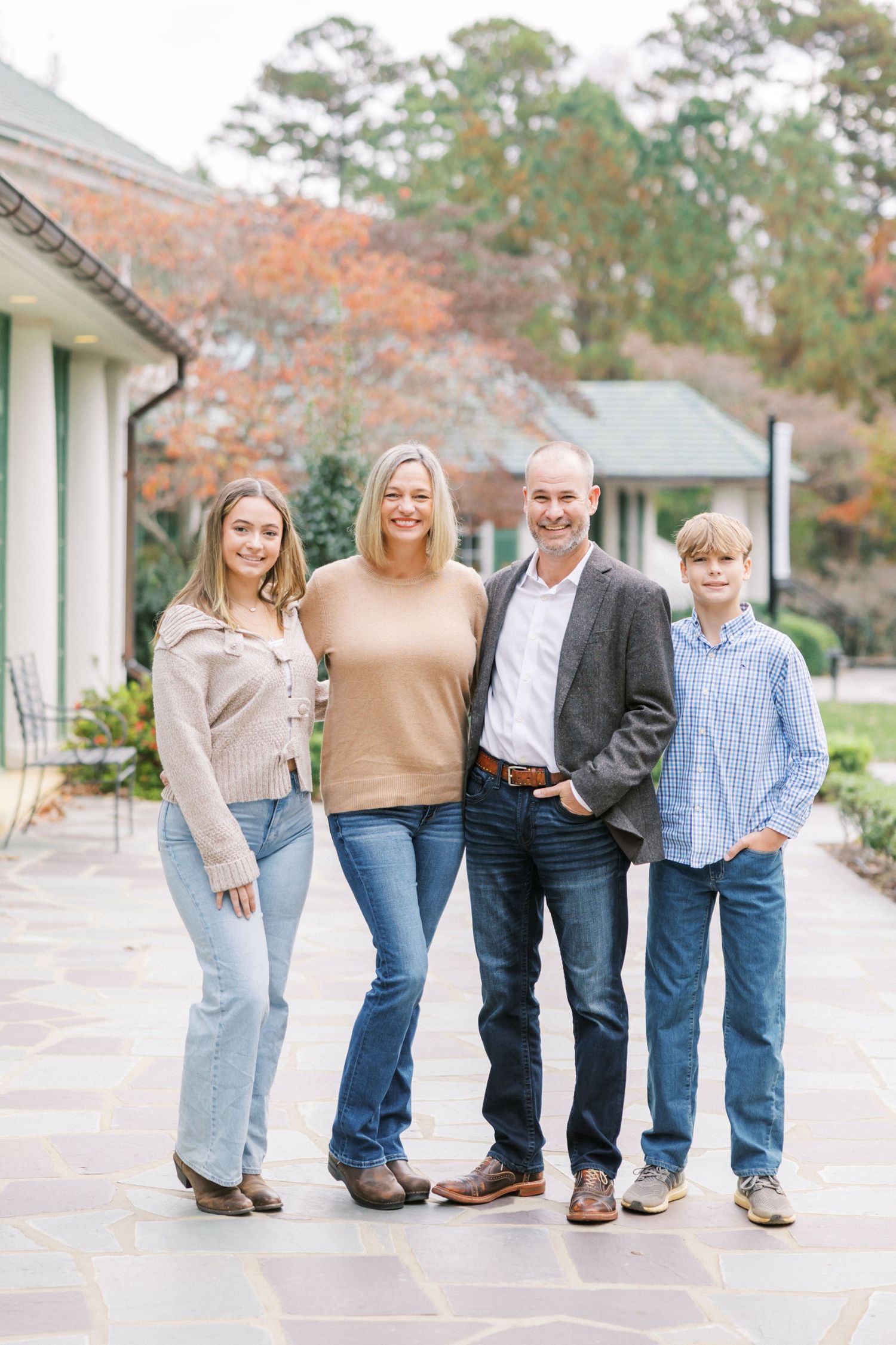 Family of four poses together on stone patio during autumn with fall foliage and house in background.