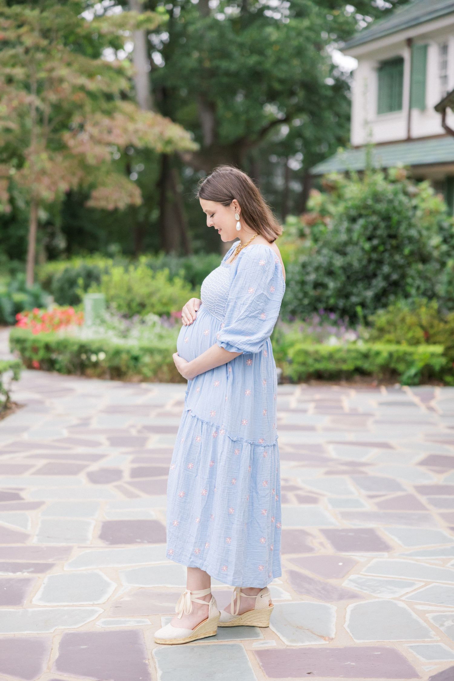 Expectant mother in light blue dress stands on stone patio in garden setting.