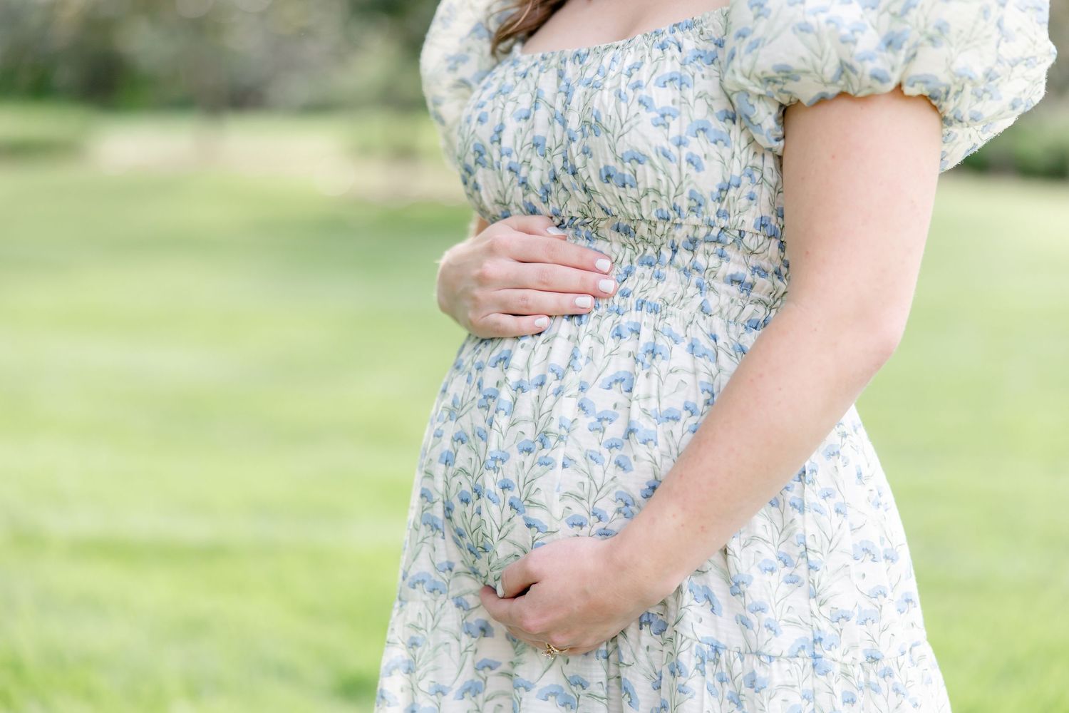 Someone wearing a floral puff sleeve dress cradles their pregnant belly while standing in a sunny garden setting.