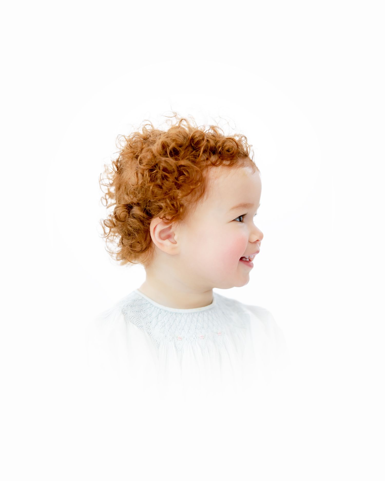 Side profile of a smiling toddler with auburn curls in a white shirt on white background.