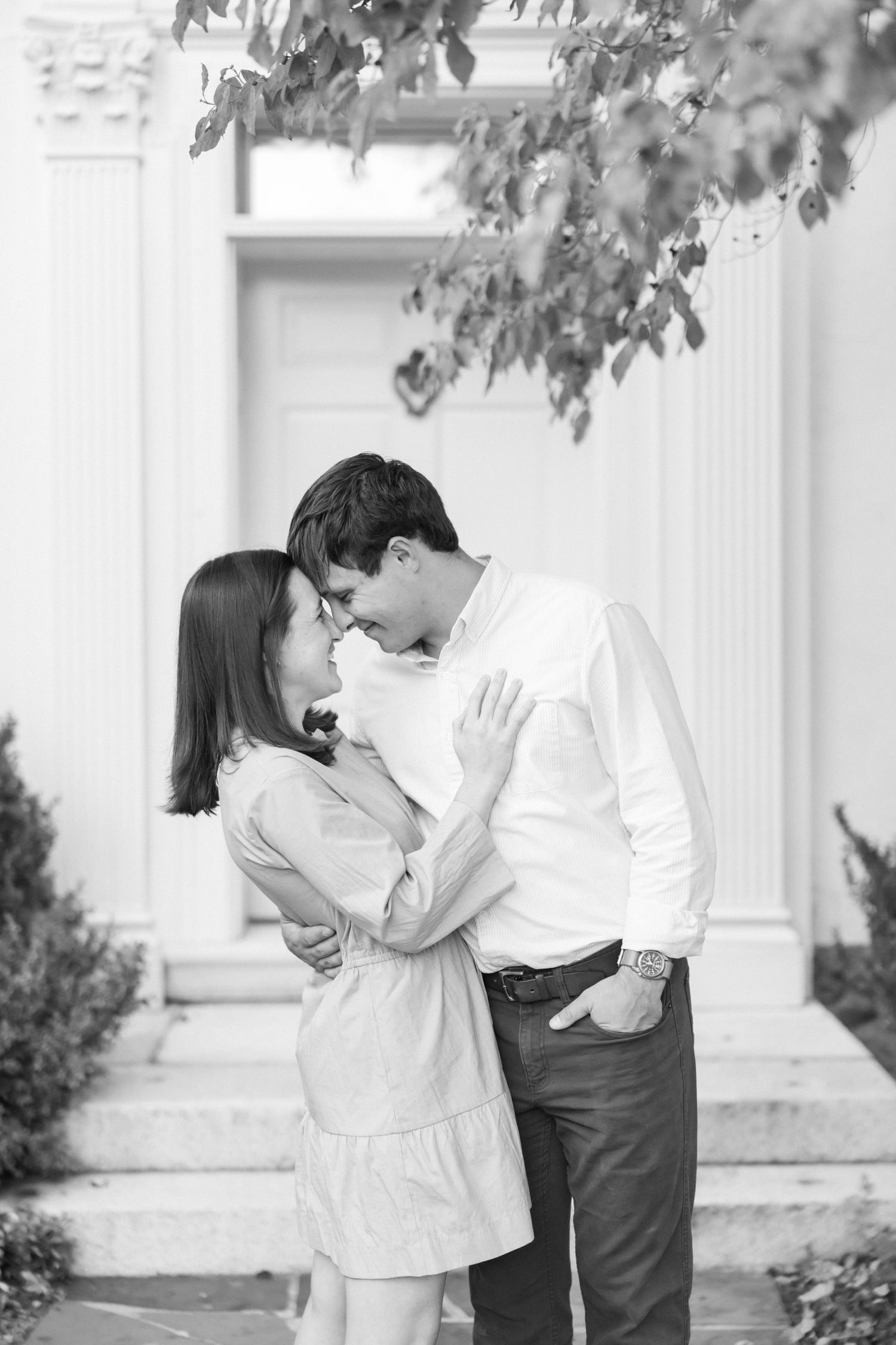 A romantic black and white photograph of a couple embracing outside a white building with foliage.