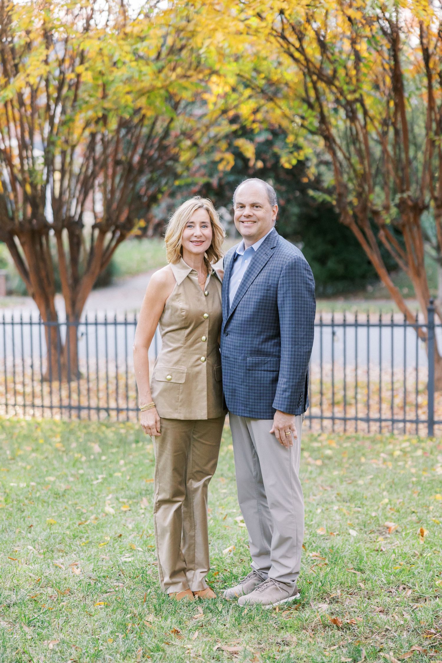 A couple poses together outdoors on a fall day with yellow autumn leaves on trees in the background.