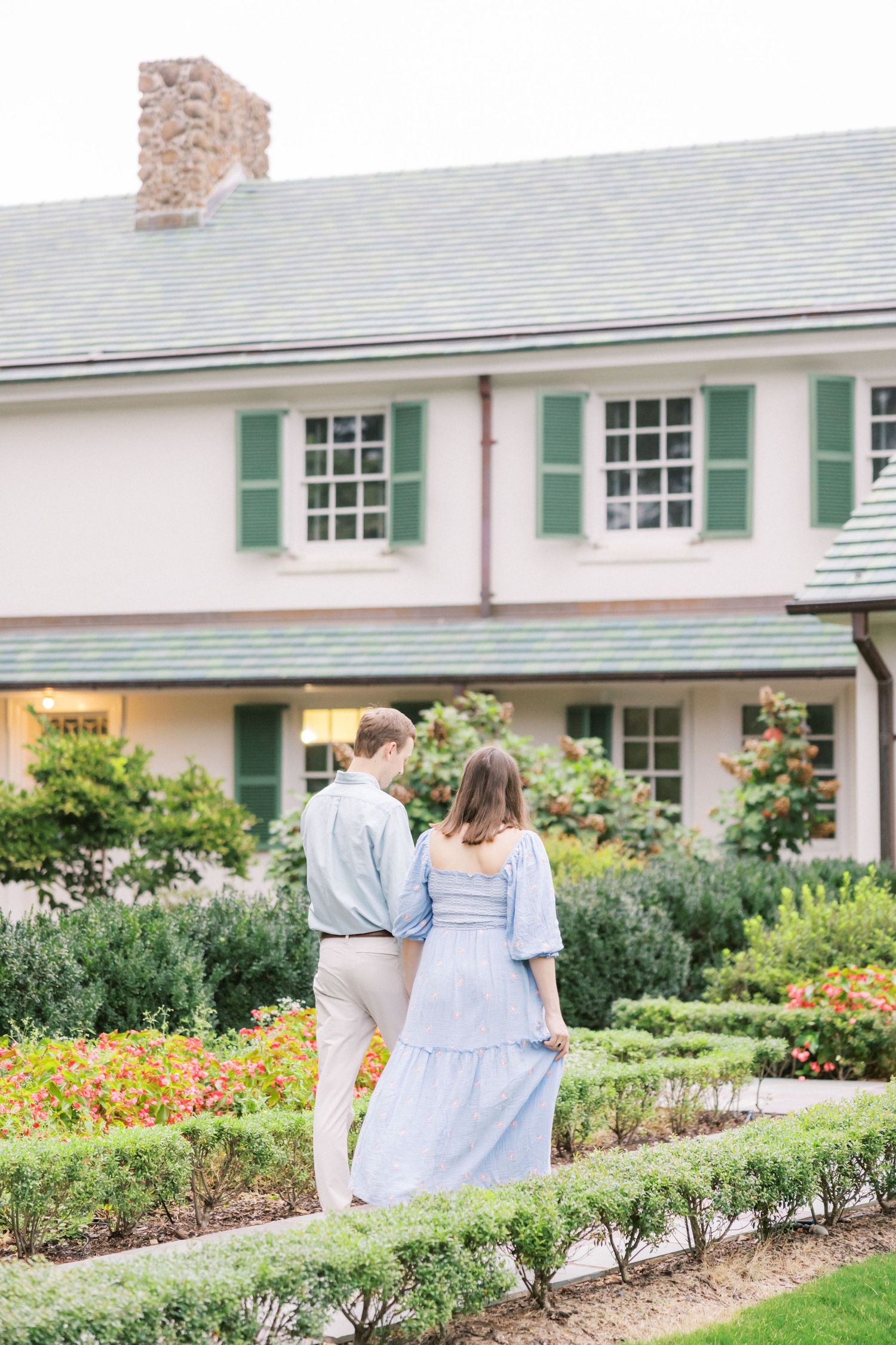 Couple stands in front of colonial style house with green shutters and colorful garden beds.