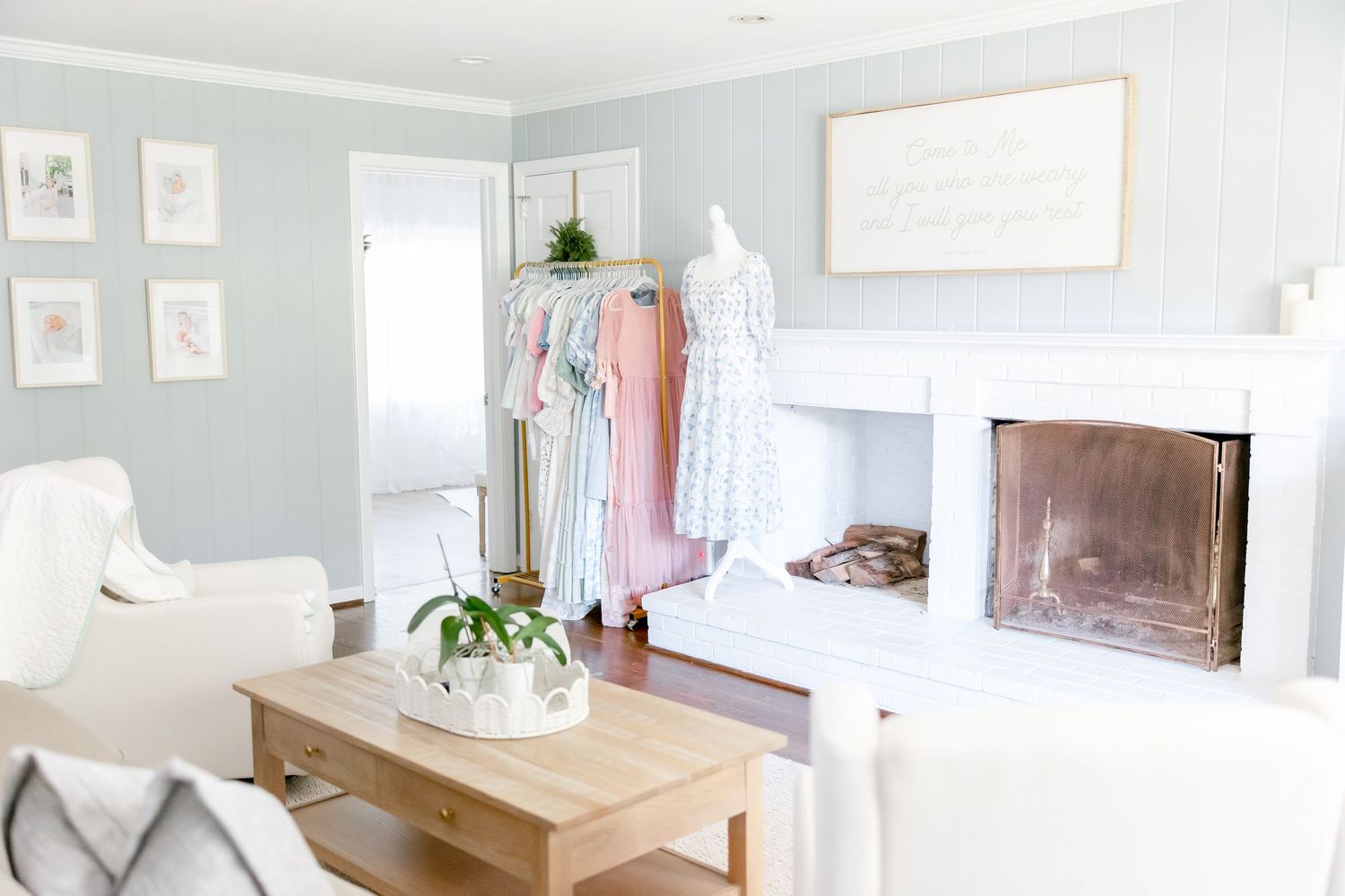 A bright minimalist living room with pale blue walls, wooden coffee table, white fireplace and clothing rack with pastel garments.