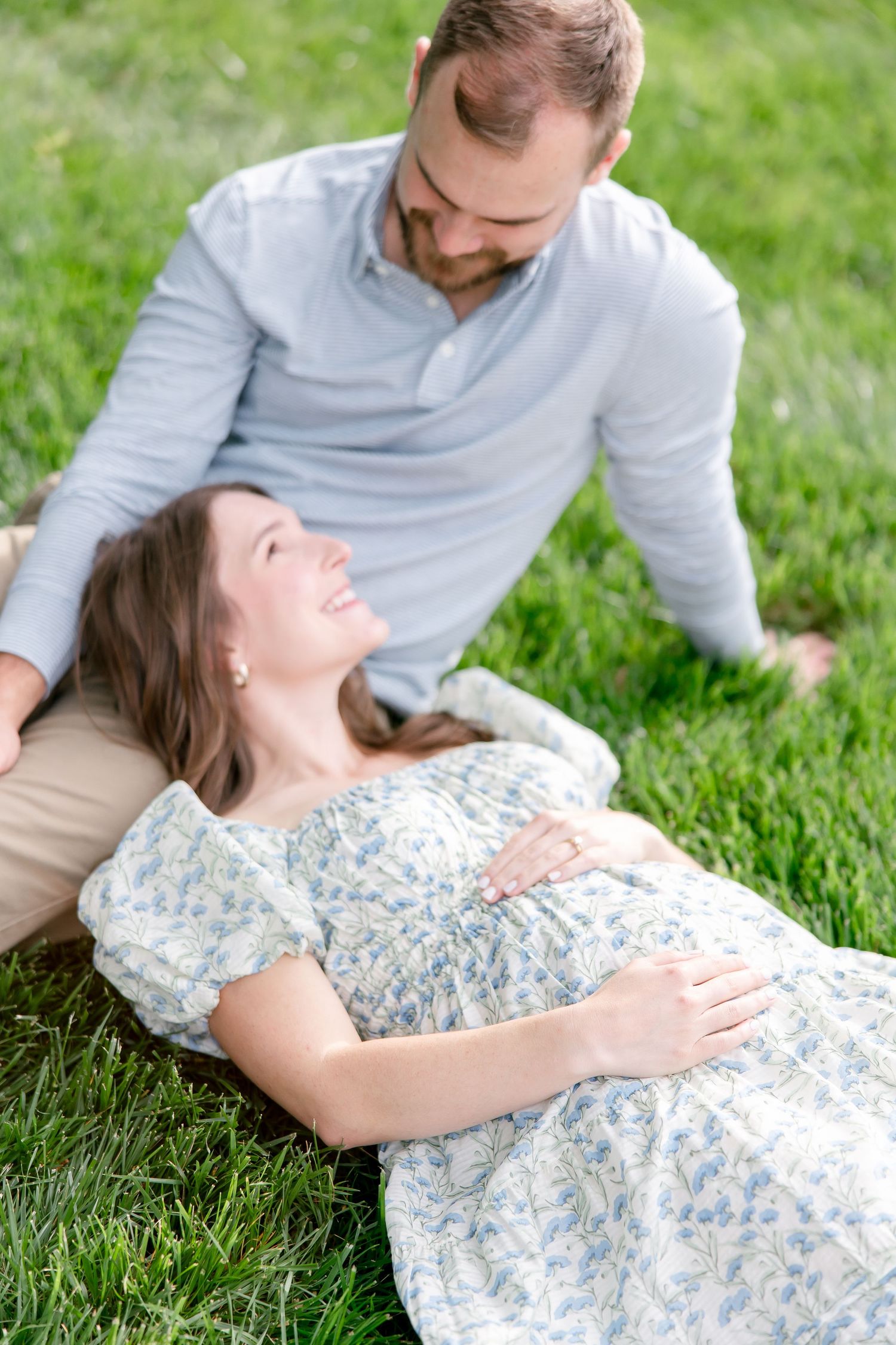 A couple relaxes together on green grass, with her wearing a floral dress and him in a light gray sweater.