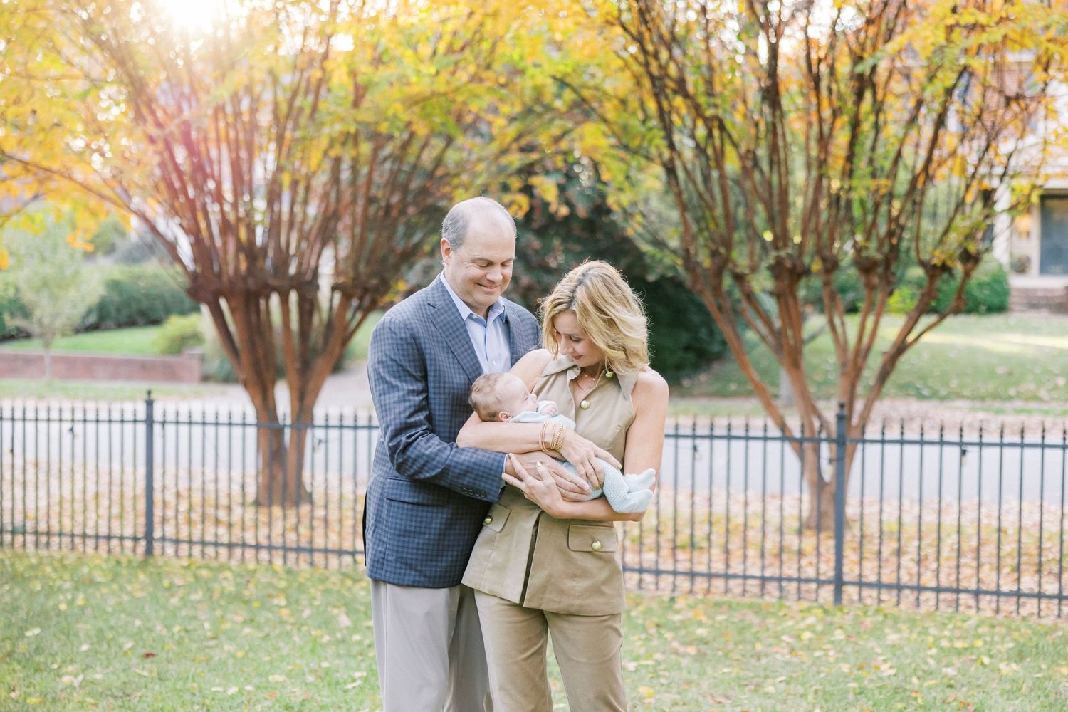 A couple embraces on a lawn during autumn with yellow trees and iron fence in the background.