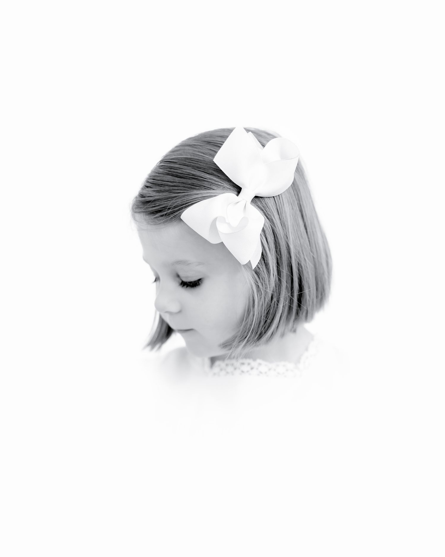 Black and white photo of a young child looking downward with a large white bow in their hair.