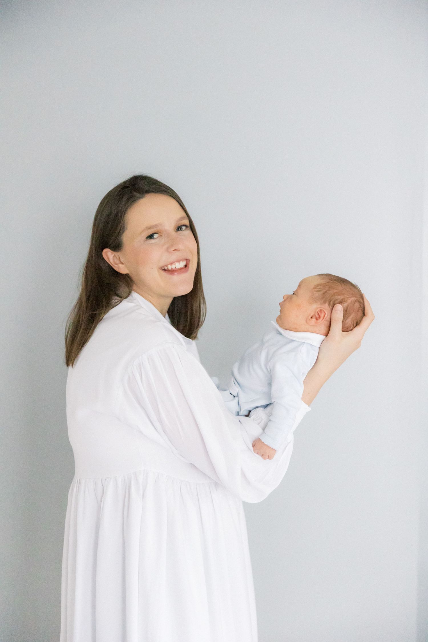 A person in a white dress holds and gazes lovingly at a newborn baby against a gray wall.