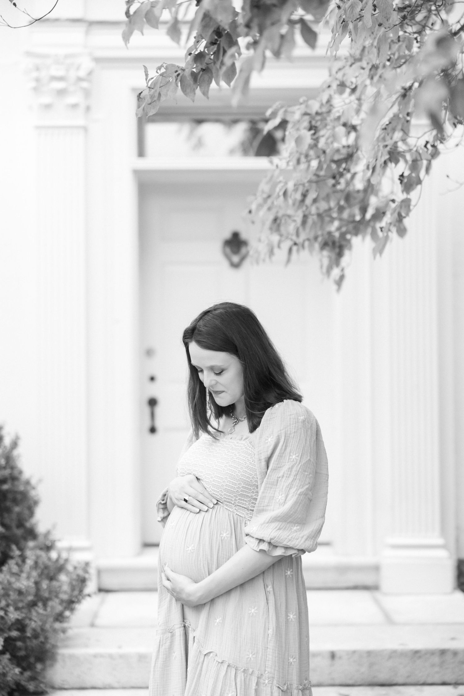 Expectant woman in flowing dress stands contemplatively by white colonial doorway with green shutters.
