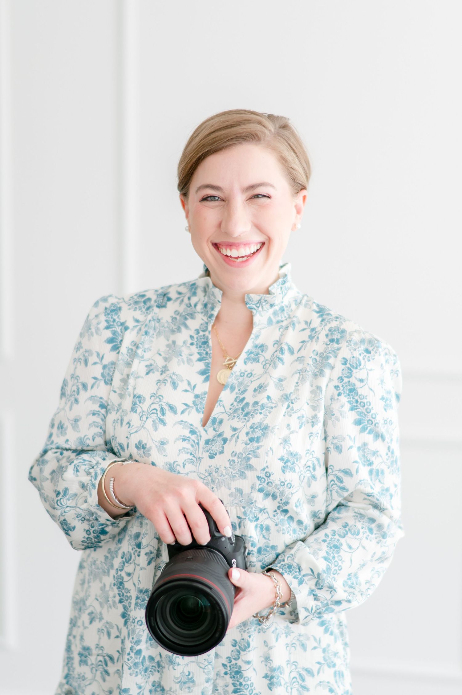 Professional photographer wearing a floral dress holding a camera with telephoto lens against white background.