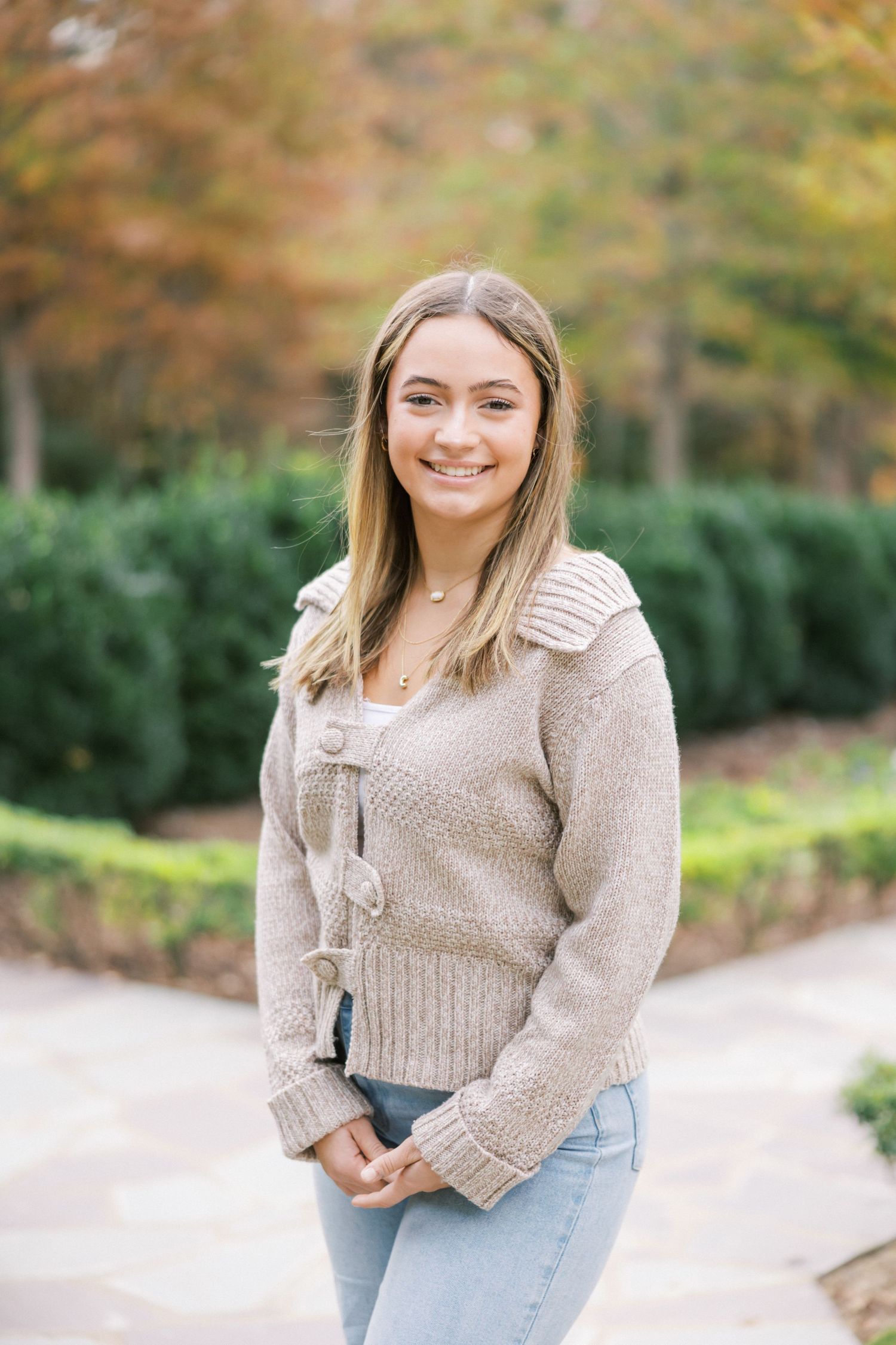 A person wearing a beige knit sweater and light jeans poses outdoors on a pathway with autumn foliage in background.