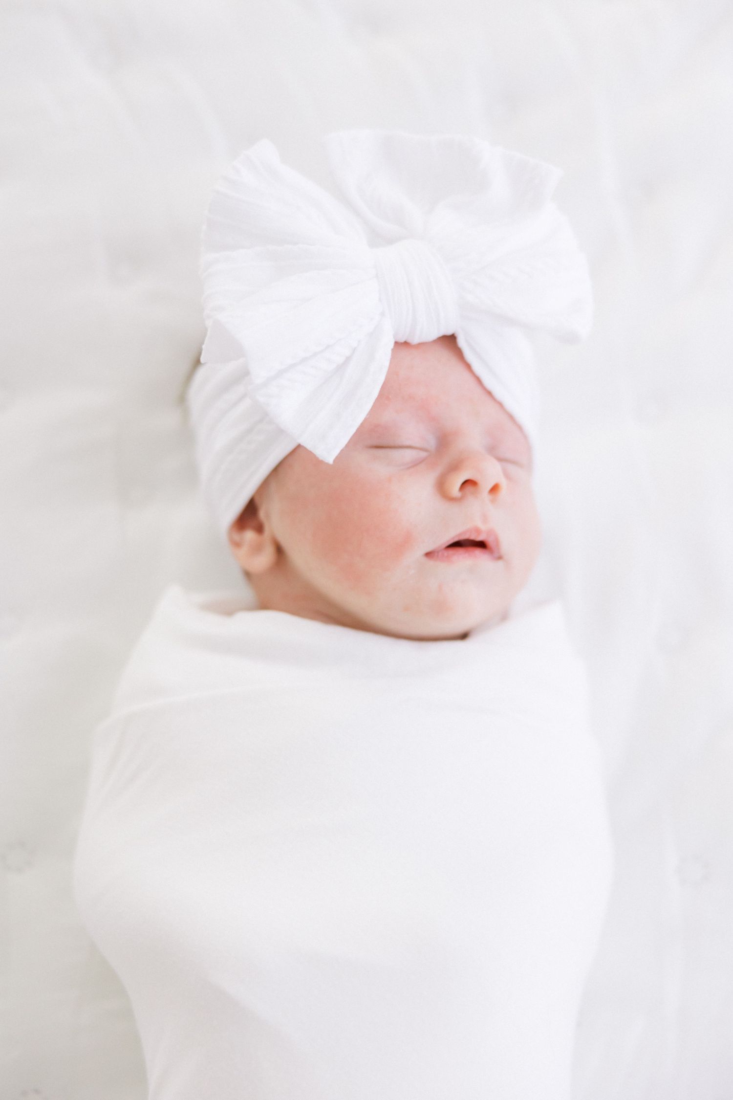Sleeping newborn wearing white bow headband and swaddle during portrait session.