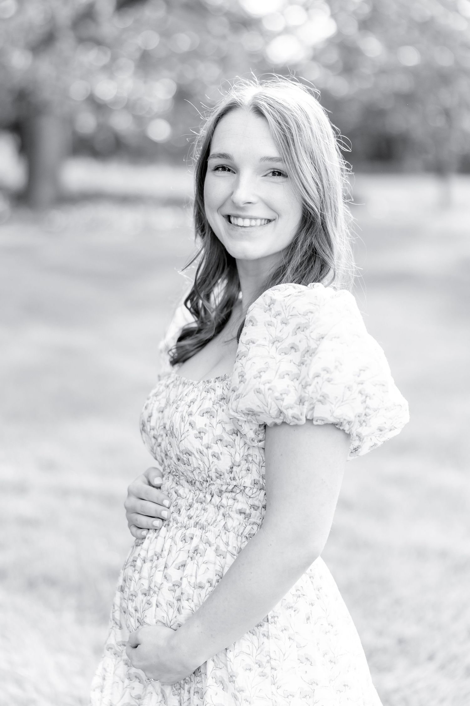 Black and white portrait of someone wearing a floral puff sleeve dress while cradling a baby bump outdoors.