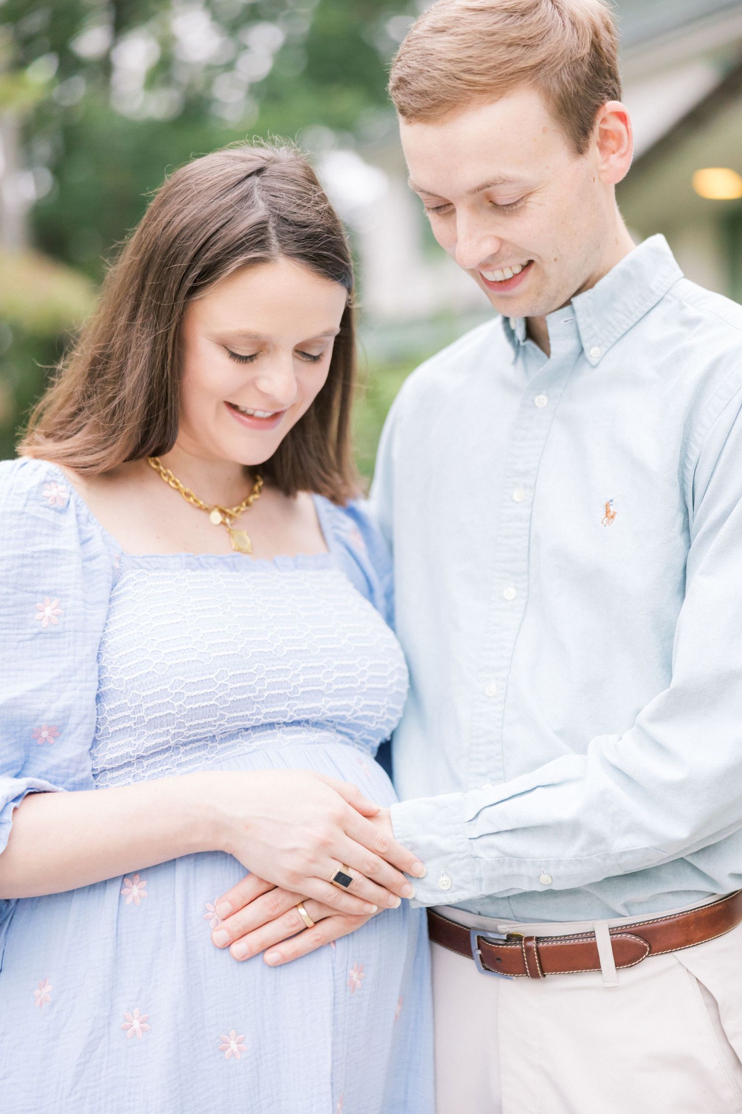A couple in light blue and white outfits share a tender moment while looking down at an engagement ring.