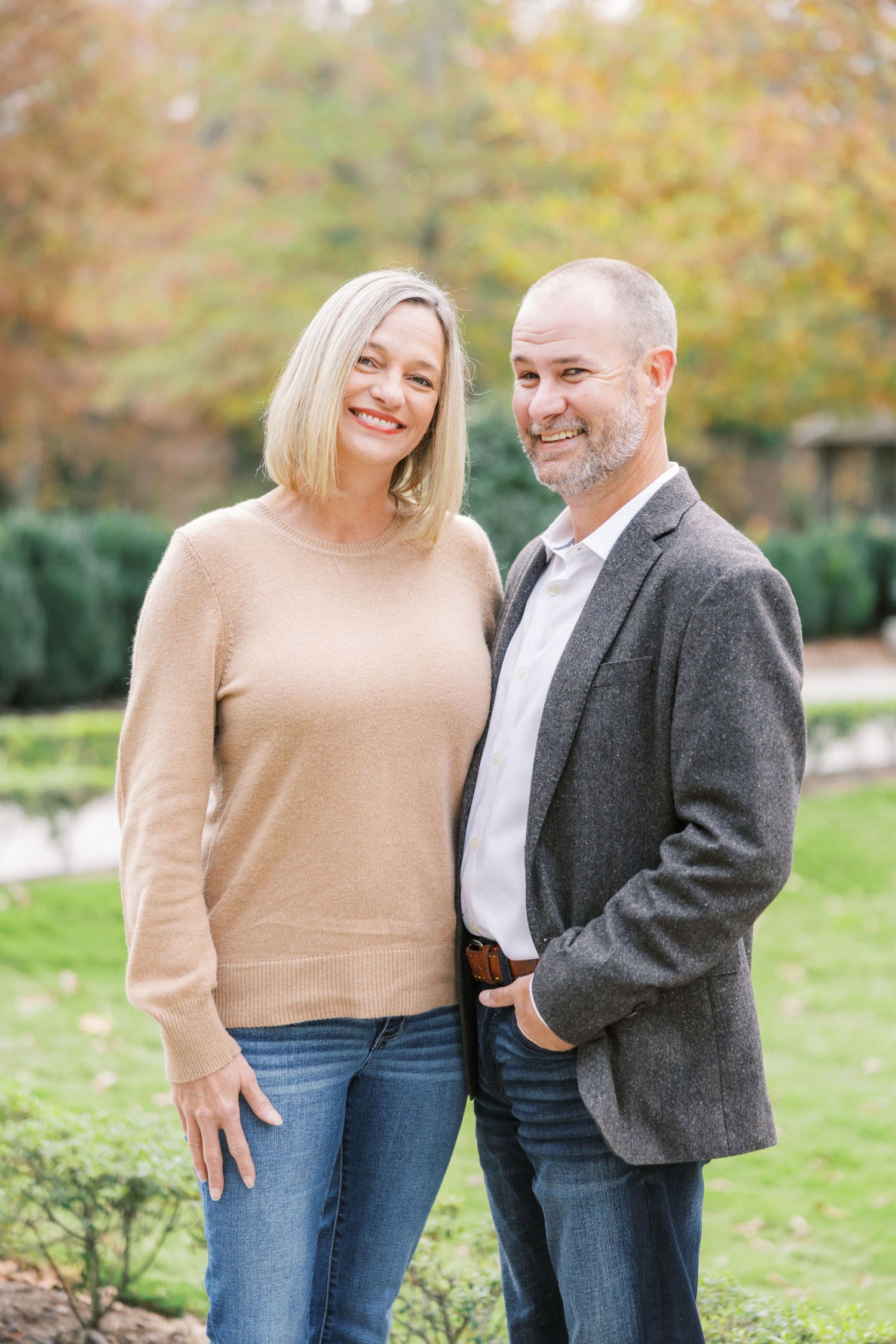 Couple dressed in casual fall attire smiling together outdoors with autumn foliage in the background.