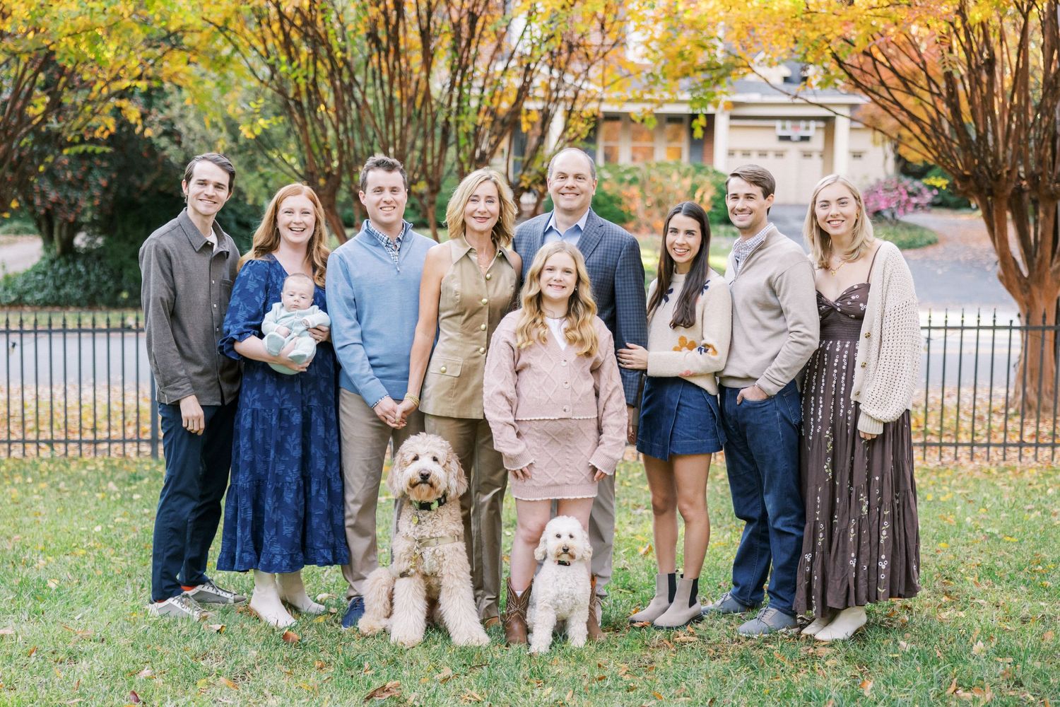 A group stands together on a lawn with two cream-colored doodle dogs in front of an iron fence on an autumn day.