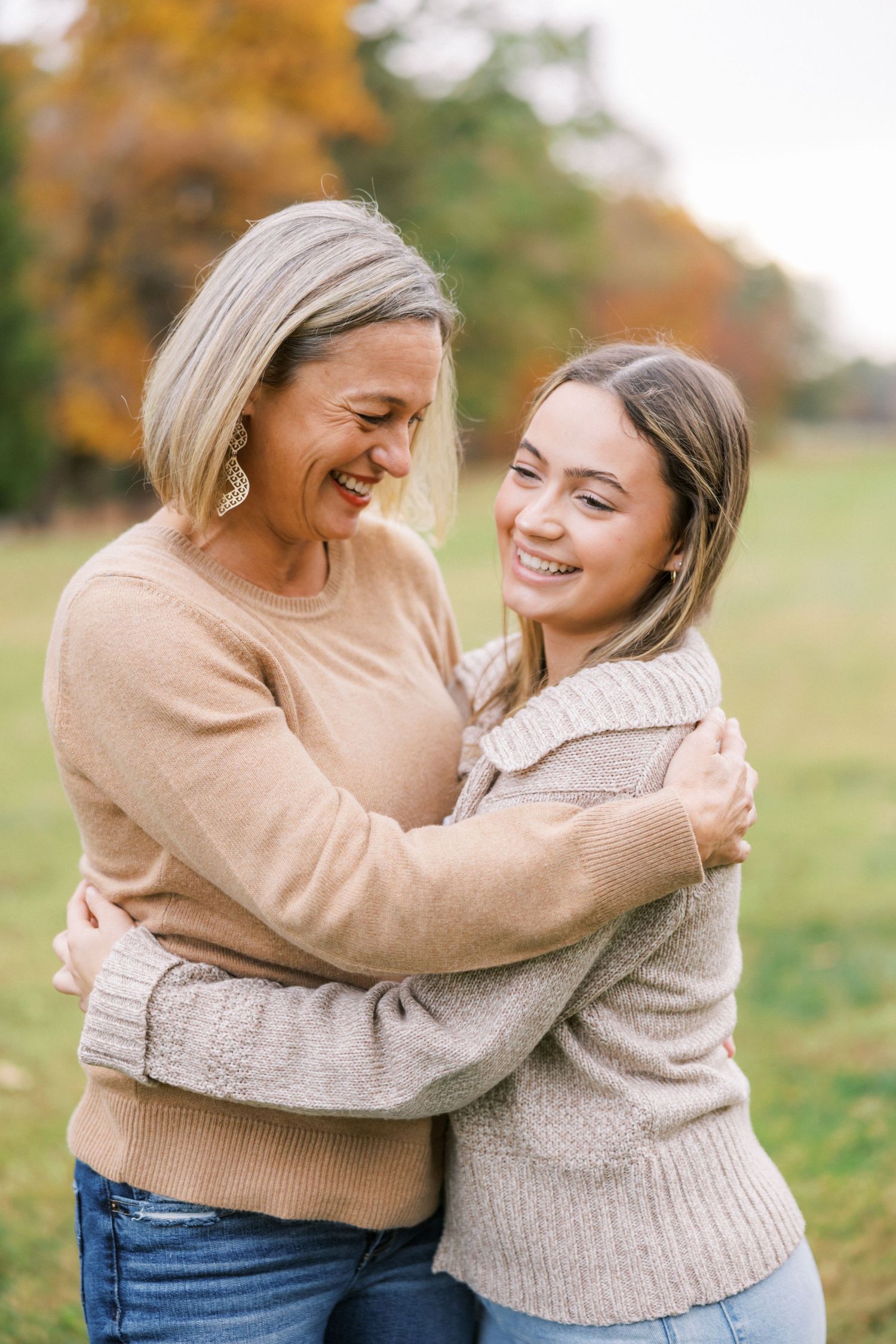 Two friends share a warm embrace and genuine laughter while standing in a park during fall season.