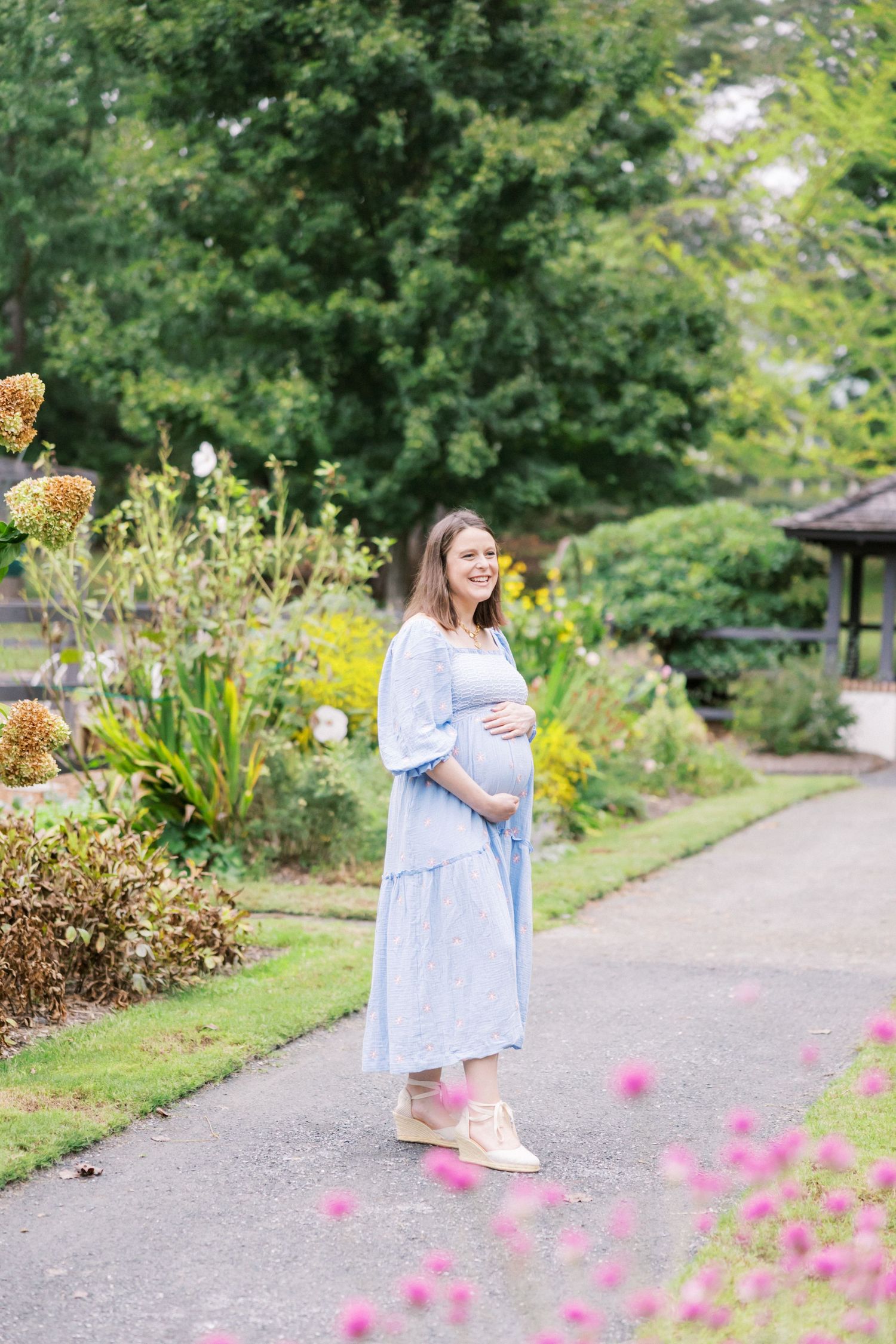 A pregnant woman in a light blue dress stands on a garden path surrounded by blooming flowers and greenery.