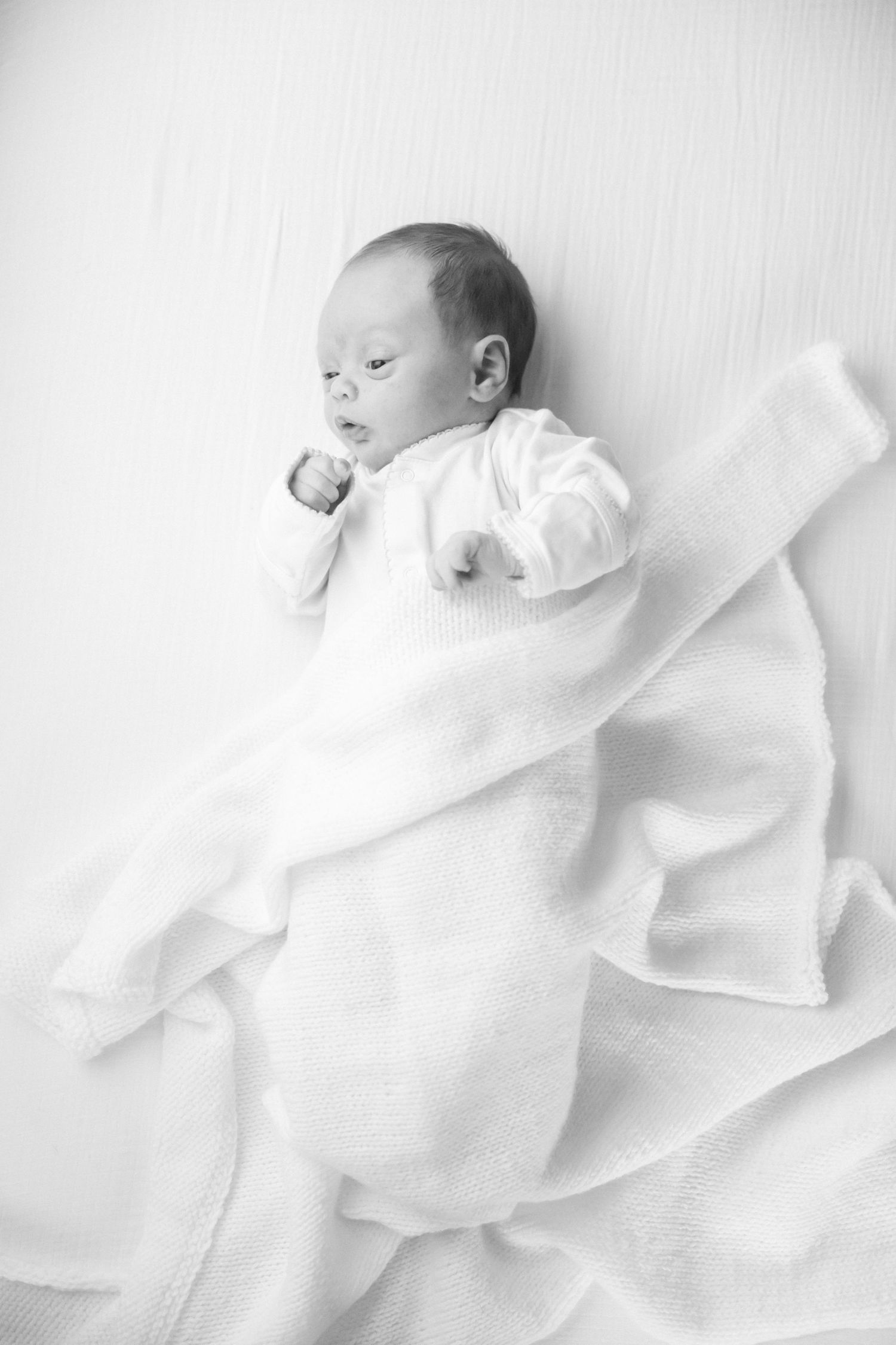 Baby in white clothing lying peacefully on soft white fabric with delicate lighting in black and white photography.